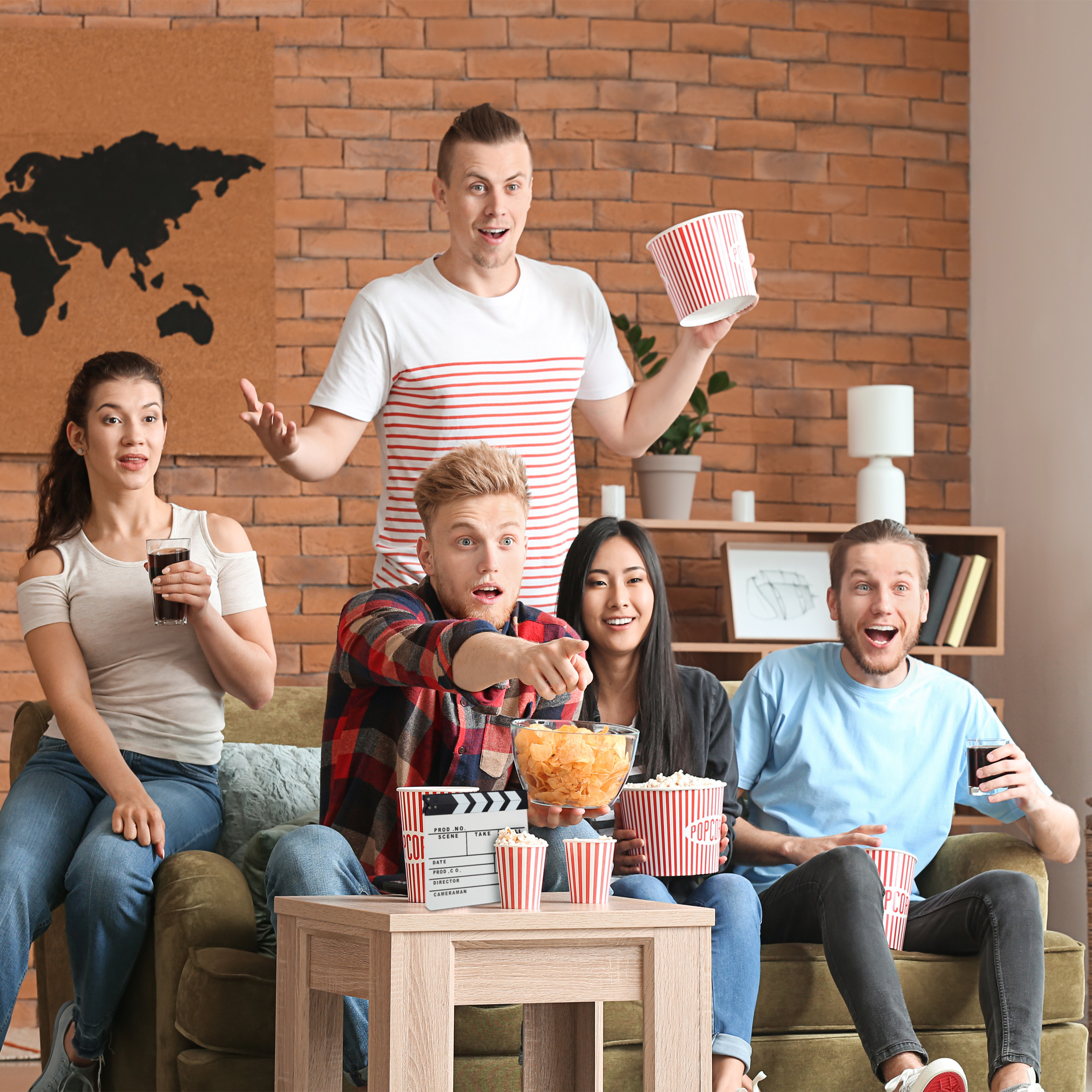 Fünf überraschte Freunde sitzen auf einem Sofa mit Popcorn und Getränken vor einer Backsteinwand.