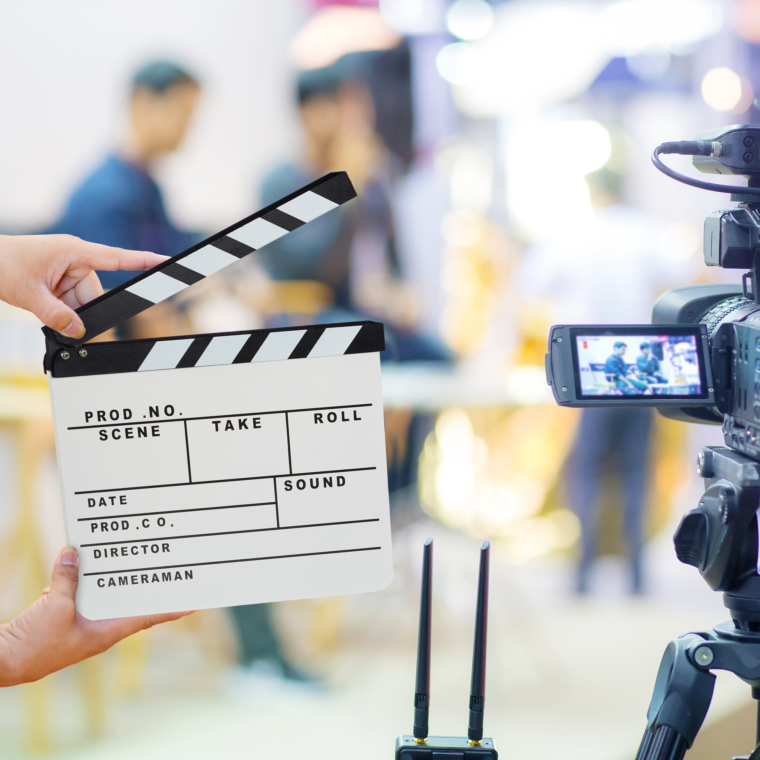 Eine Hand hält eine Filmklappe vor eine Kamera, im Hintergrund ist ein unscharfes Filmset zu sehen.