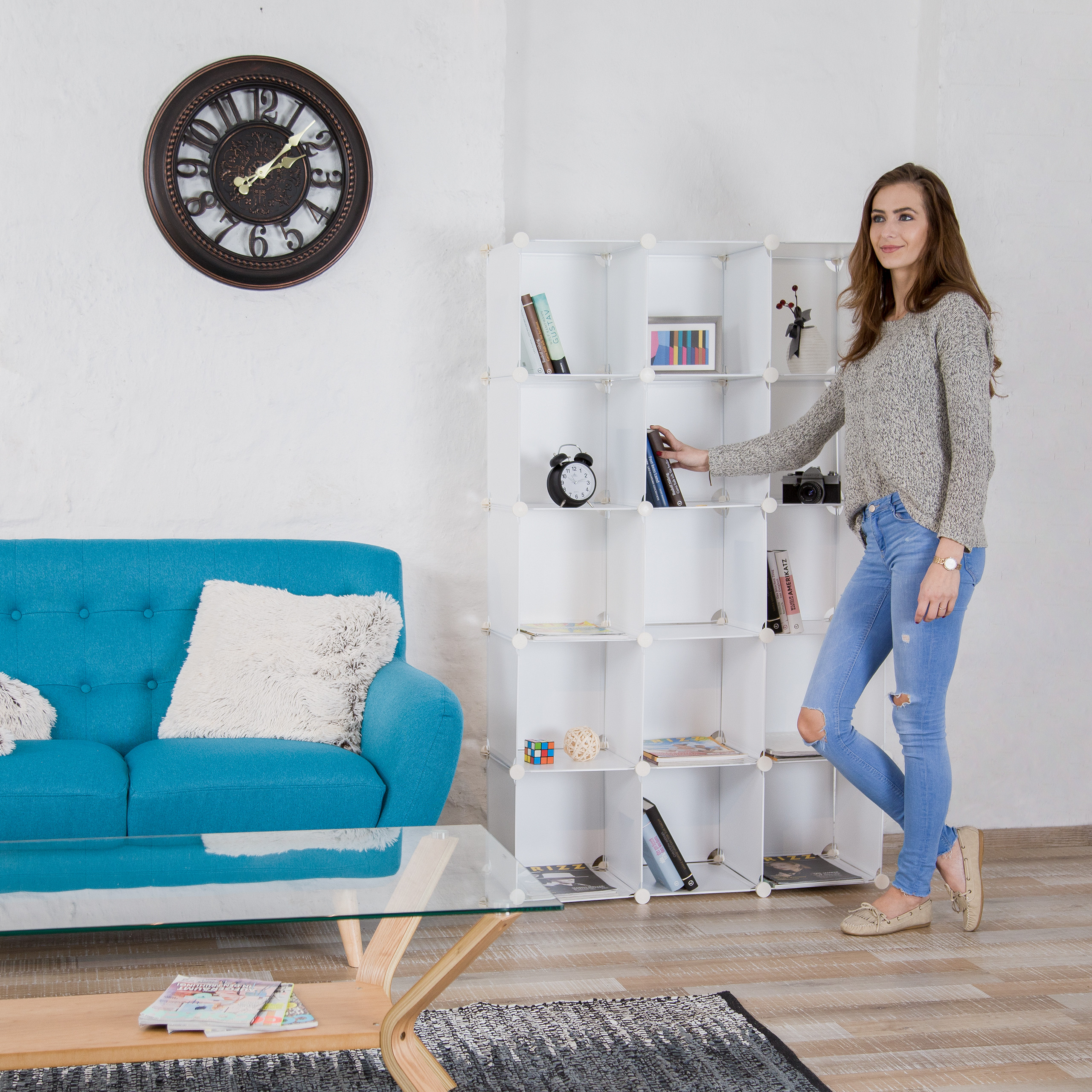 Eine Frau in Jeans und Pullover steht neben einem weißen Regal in einem modernen Wohnzimmer mit blauem Sofa.