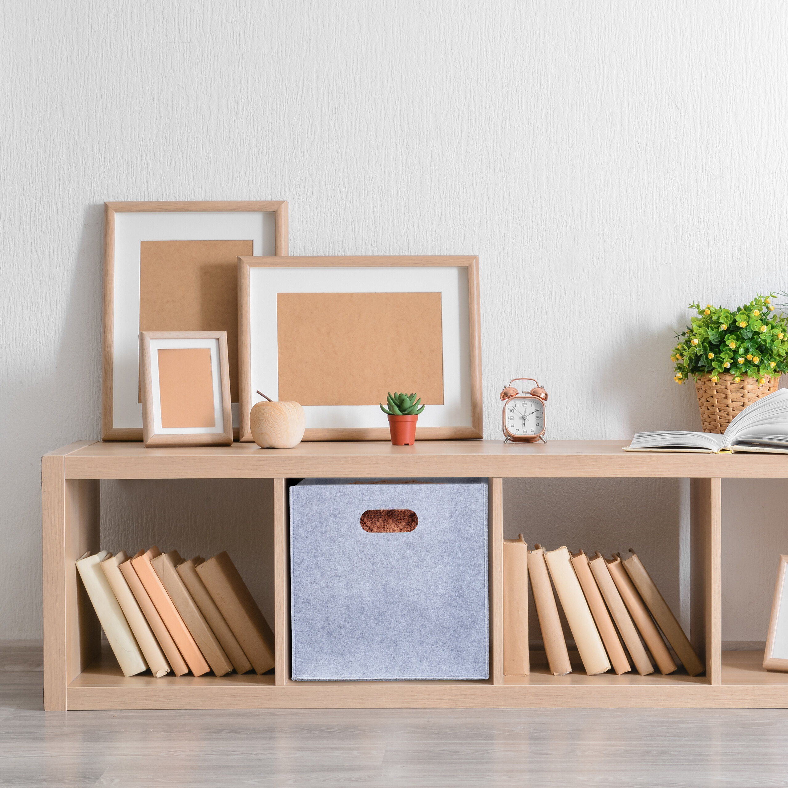 Ein Holzregal an einer weißen Wand mit Bilderrahmen, einer Uhr, Büchern, einer Aufbewahrungsbox und einer Topfpflanze.