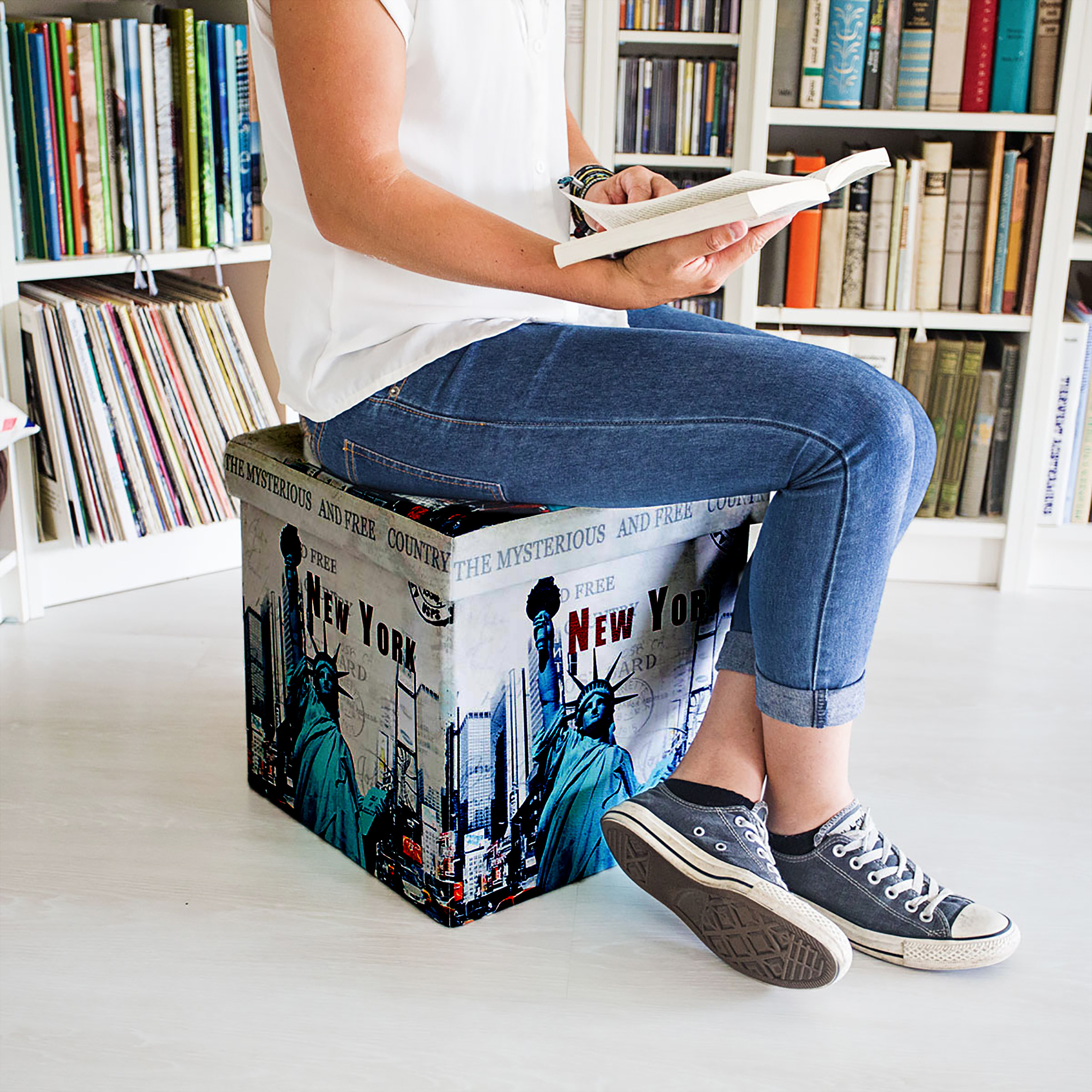Eine Person in Jeans und Turnschuhen liest ein Buch auf einer New-York-Sitzkiste vor einem Bücherregal.
