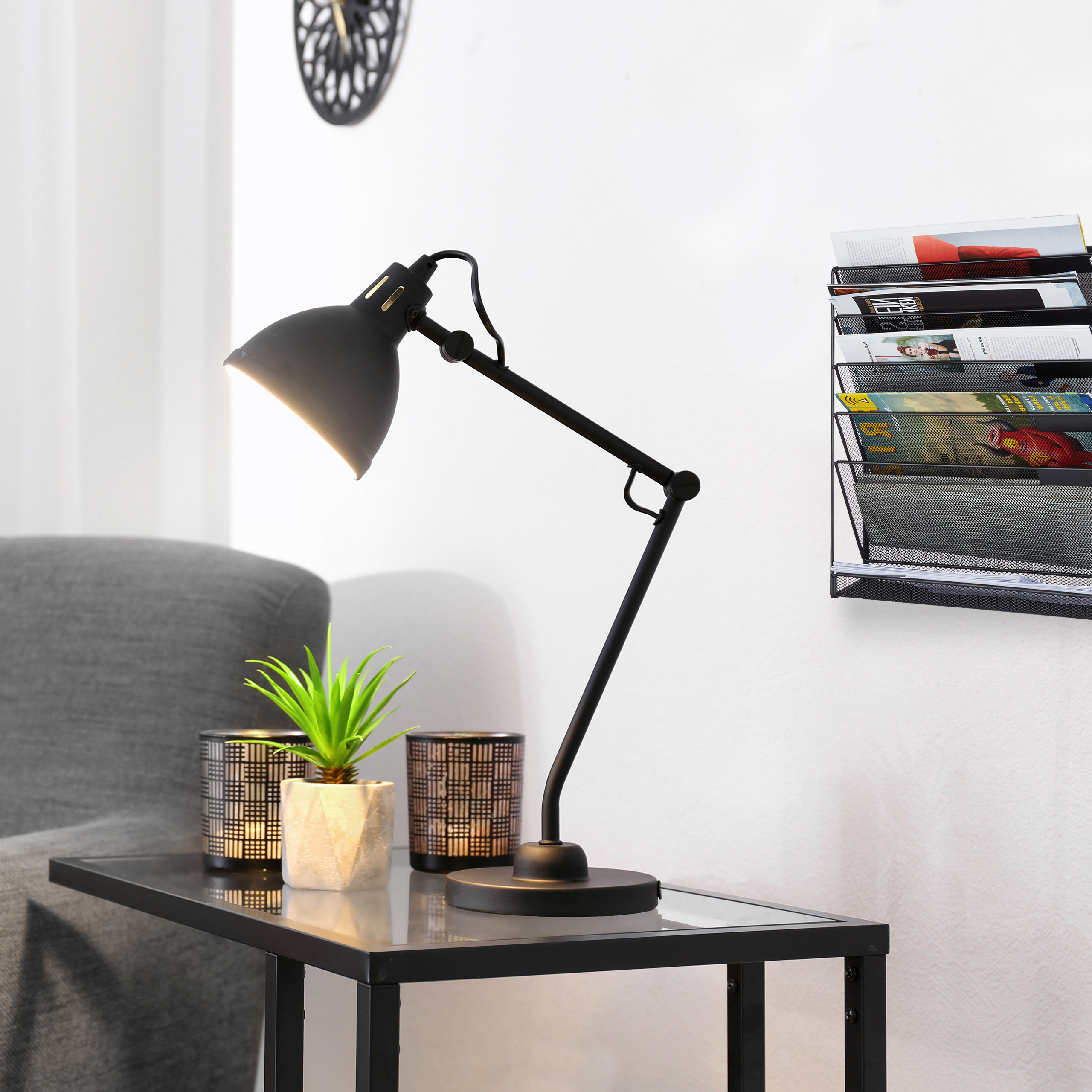 Eine schwarze Schreibtischlampe, eine kleine Pflanze und Kerzen auf einem Glastisch vor einer weißen Wand mit Zeitschriftenhalter.