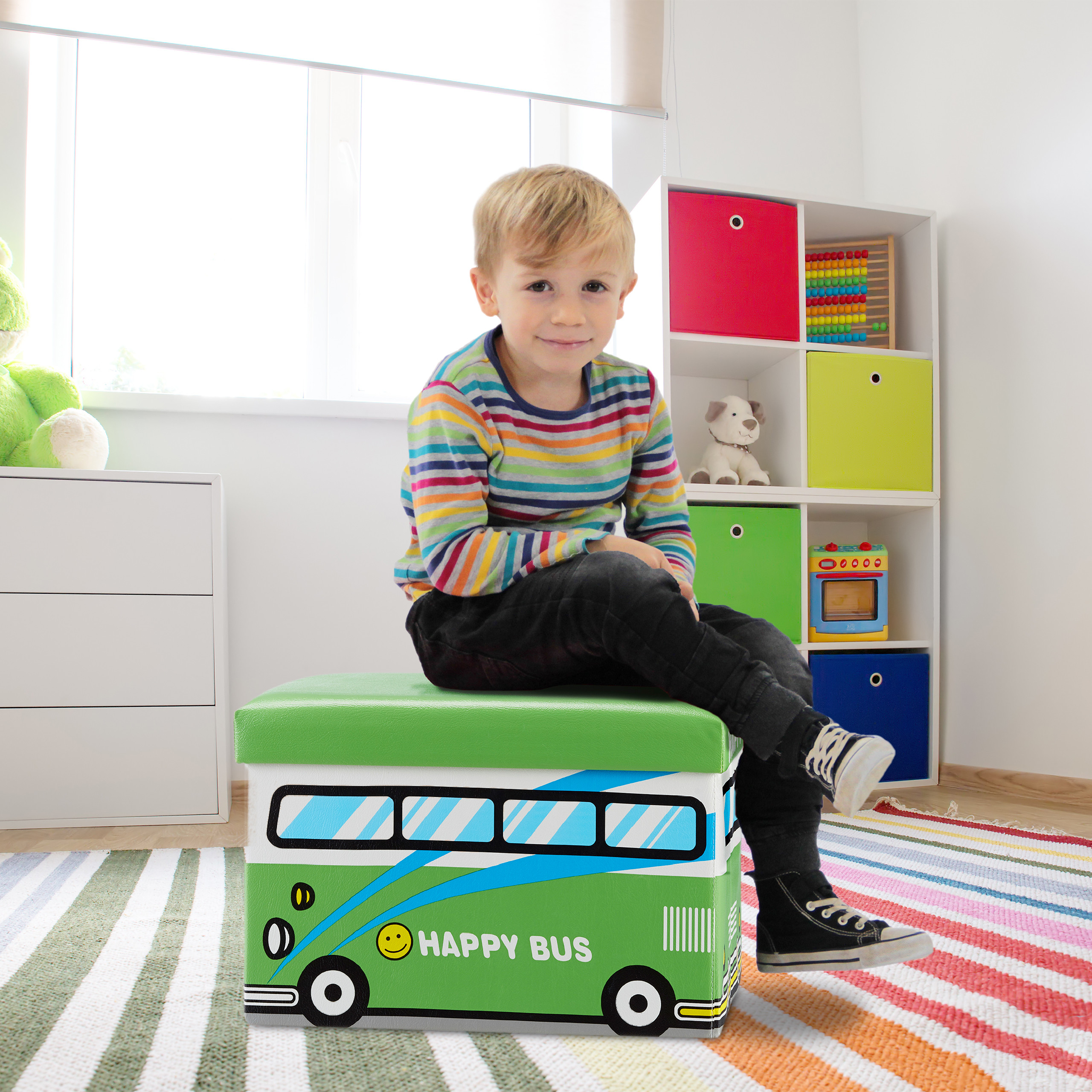 Blonde Junge im gestreiften Shirt sitzt auf einer grünen Happy-Bus-Spielzeugbox im Kinderzimmer.