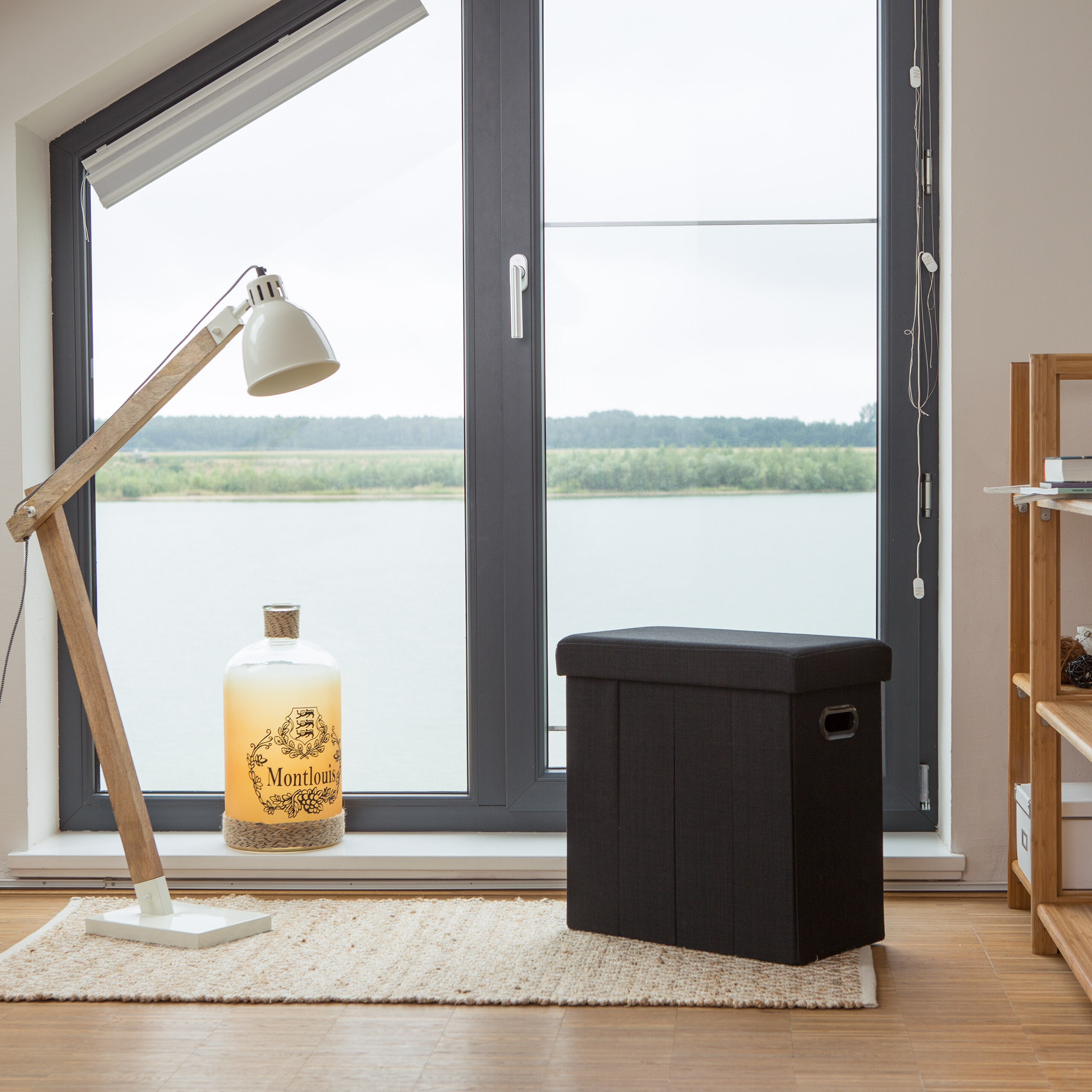 Holzlampe, dekorierte Flasche und schwarzer Hocker stehen auf einem Teppich vor einem großen Fenster mit Seeblick.