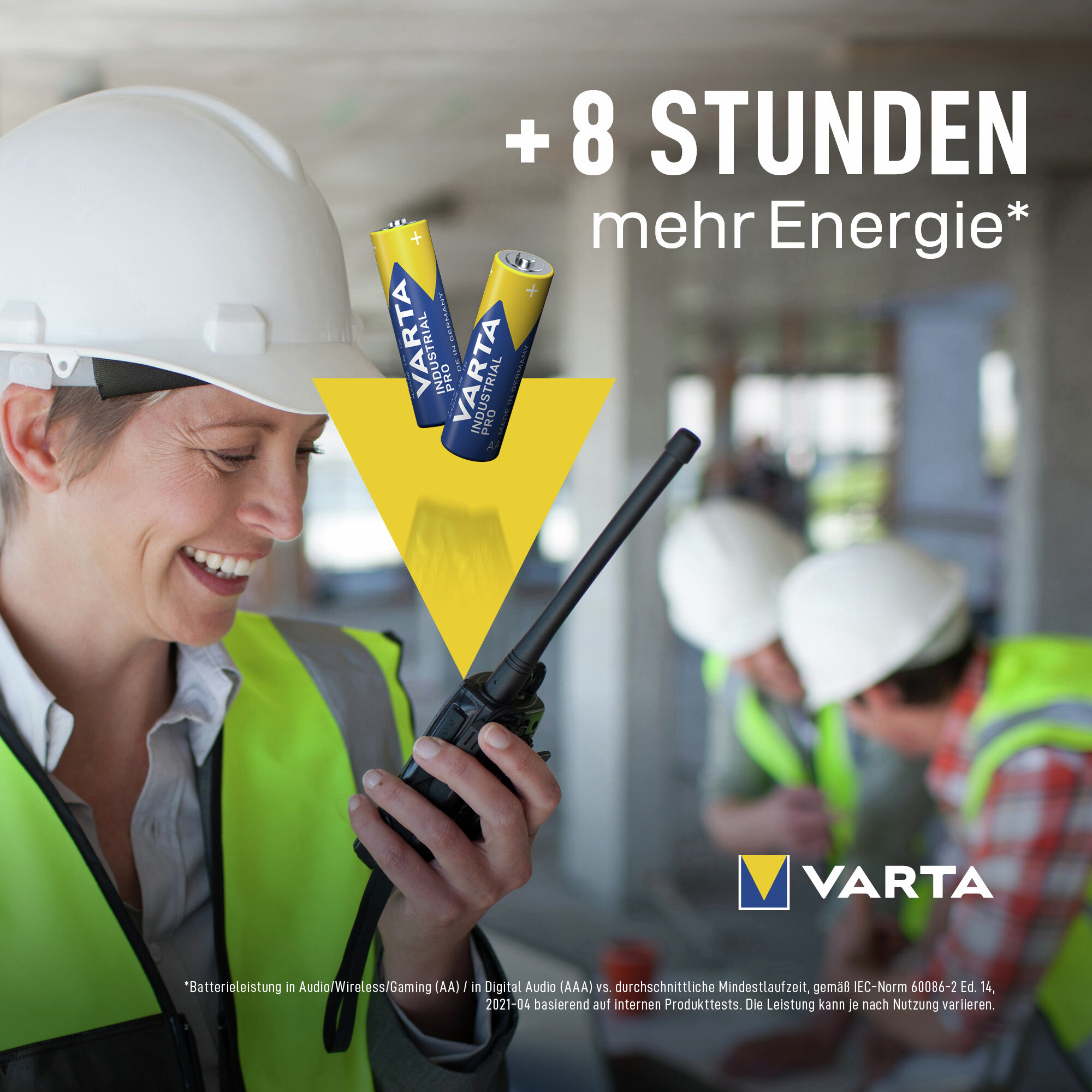 Bauarbeiterin mit Helm hält ein Funkgerät. Im Hintergrund zwei weitere Arbeiter und Varta-Akkus.
