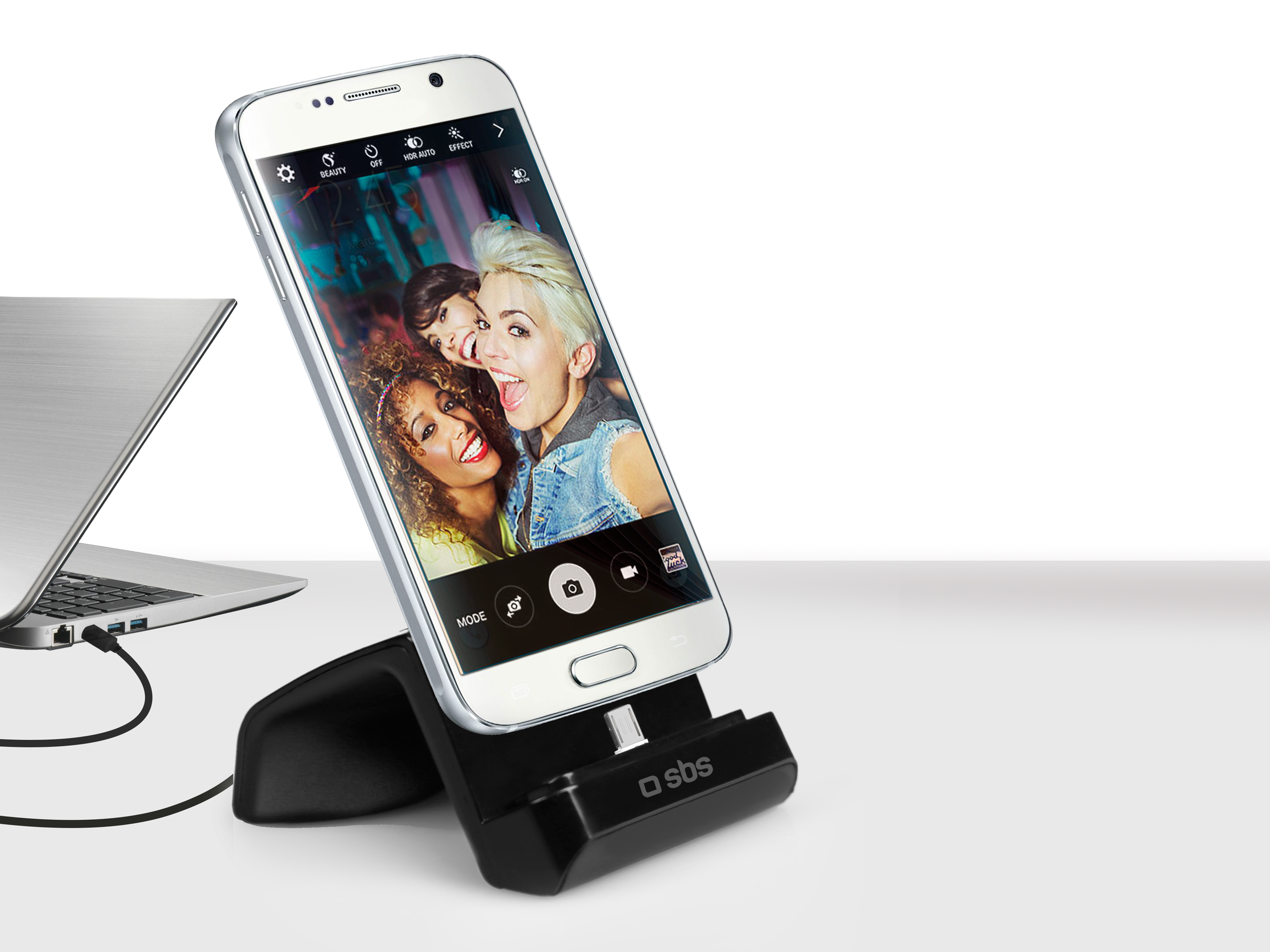 Un teléfono blanco en una base de carga negra, conectado a un portátil plateado.