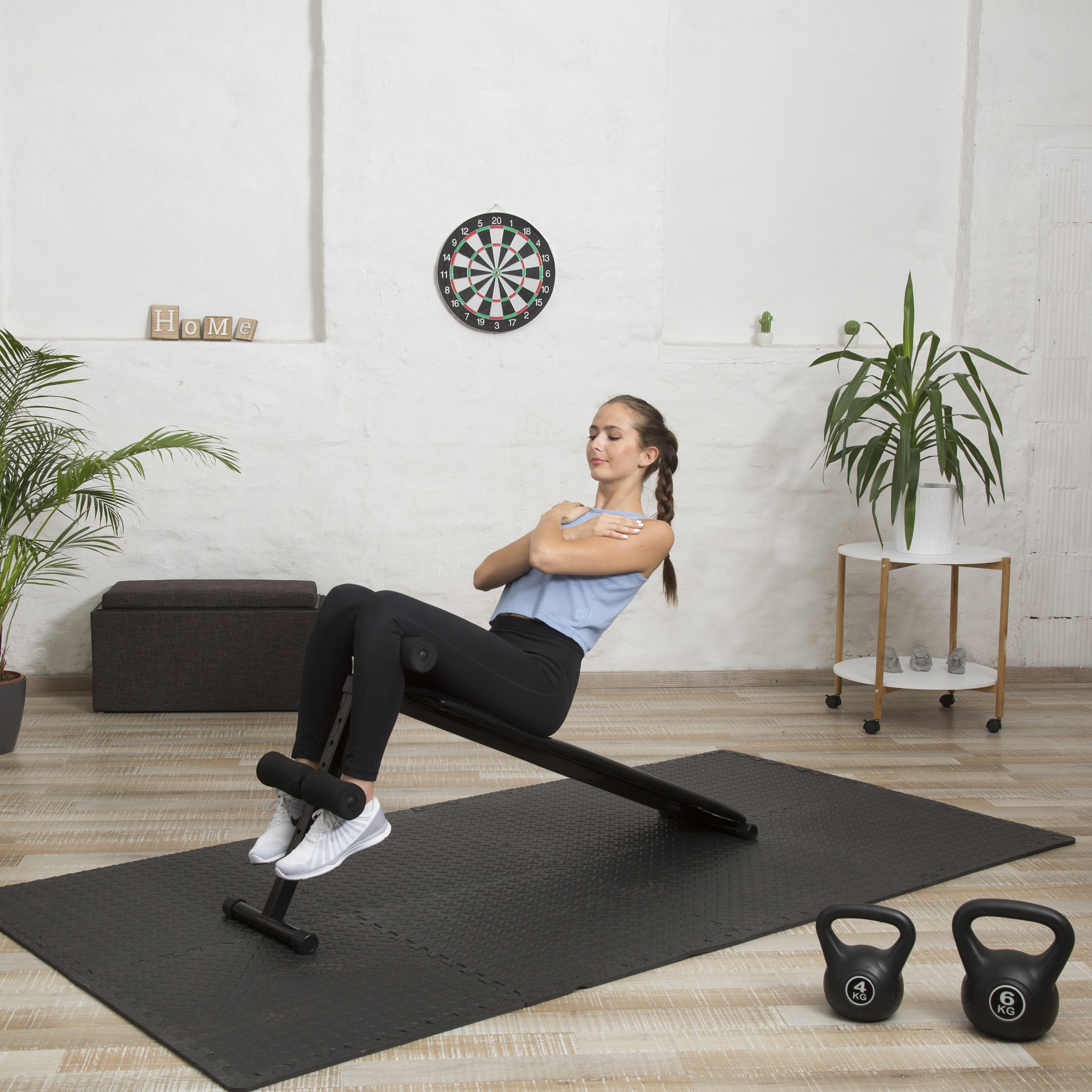 Eine Frau in Sportkleidung macht Sit-ups auf einer Bank im Wohnzimmer, vor einer weißen Wand mit Darts-Scheibe.