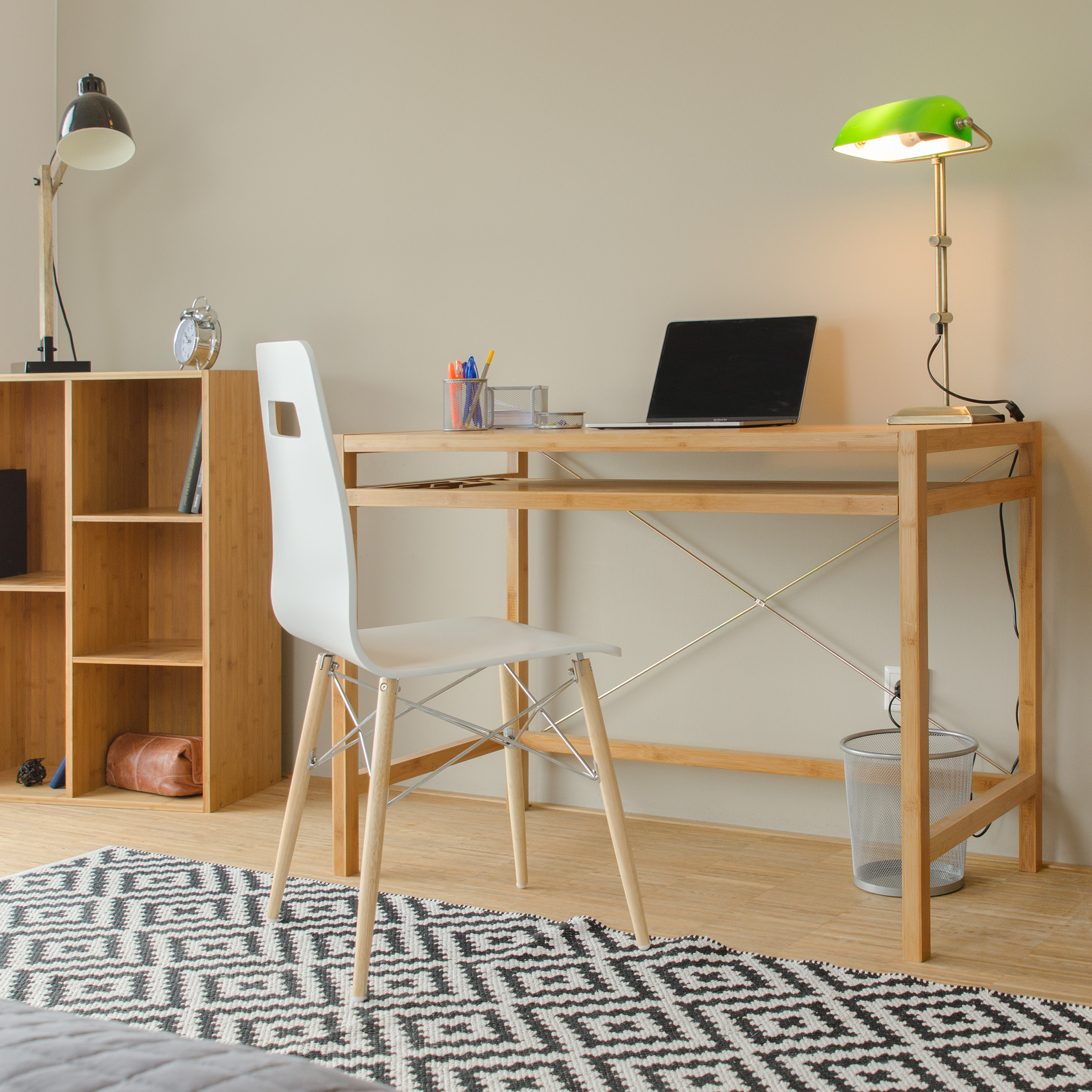 Ein Holztisch mit Laptop, grüner Lampe, weißem Stuhl und schwarz-weiß gemustertem Teppich im Vordergrund.