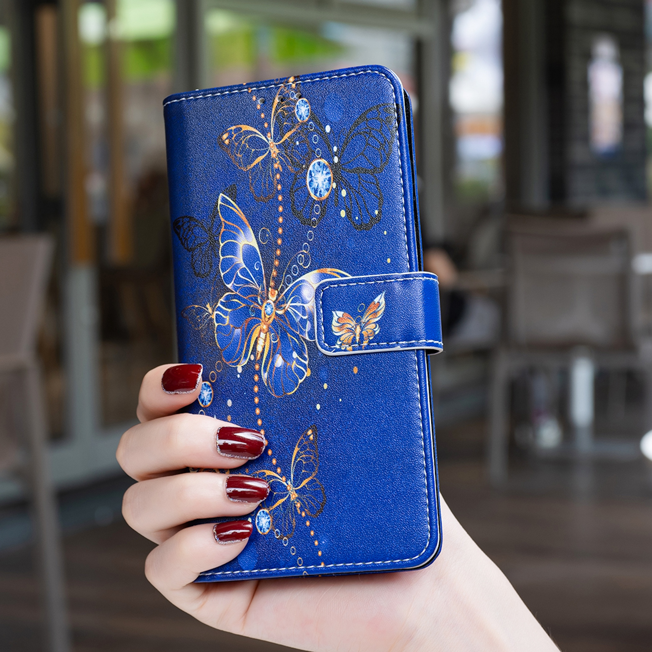 Eine Hand hält eine blaue Leder-Handyhülle mit goldenen Schmetterlings- und Juwelenmotiven.