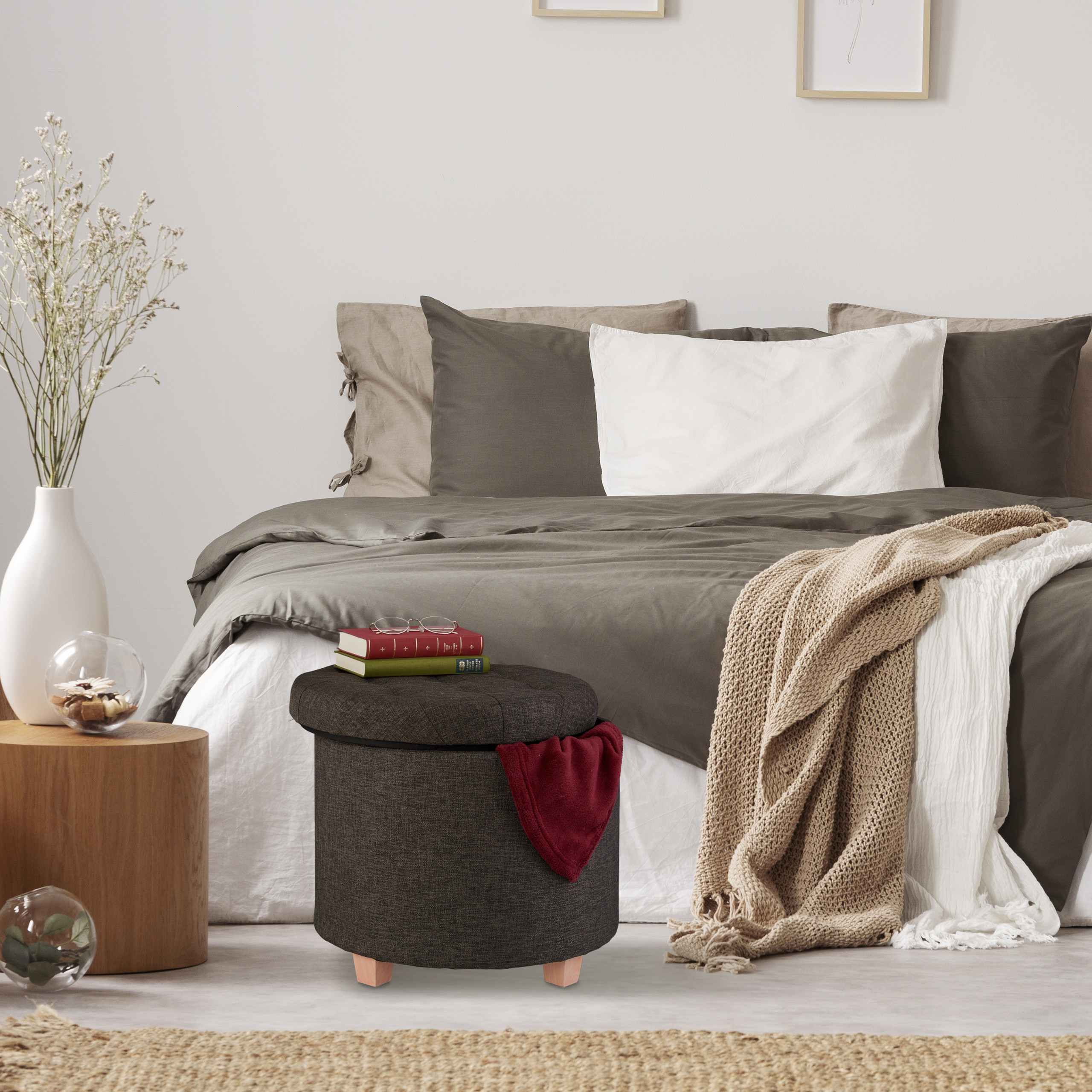 Ein gemütliches Schlafzimmer mit grauem Bett, Kissen, einer Decke und einem Hocker mit Büchern.