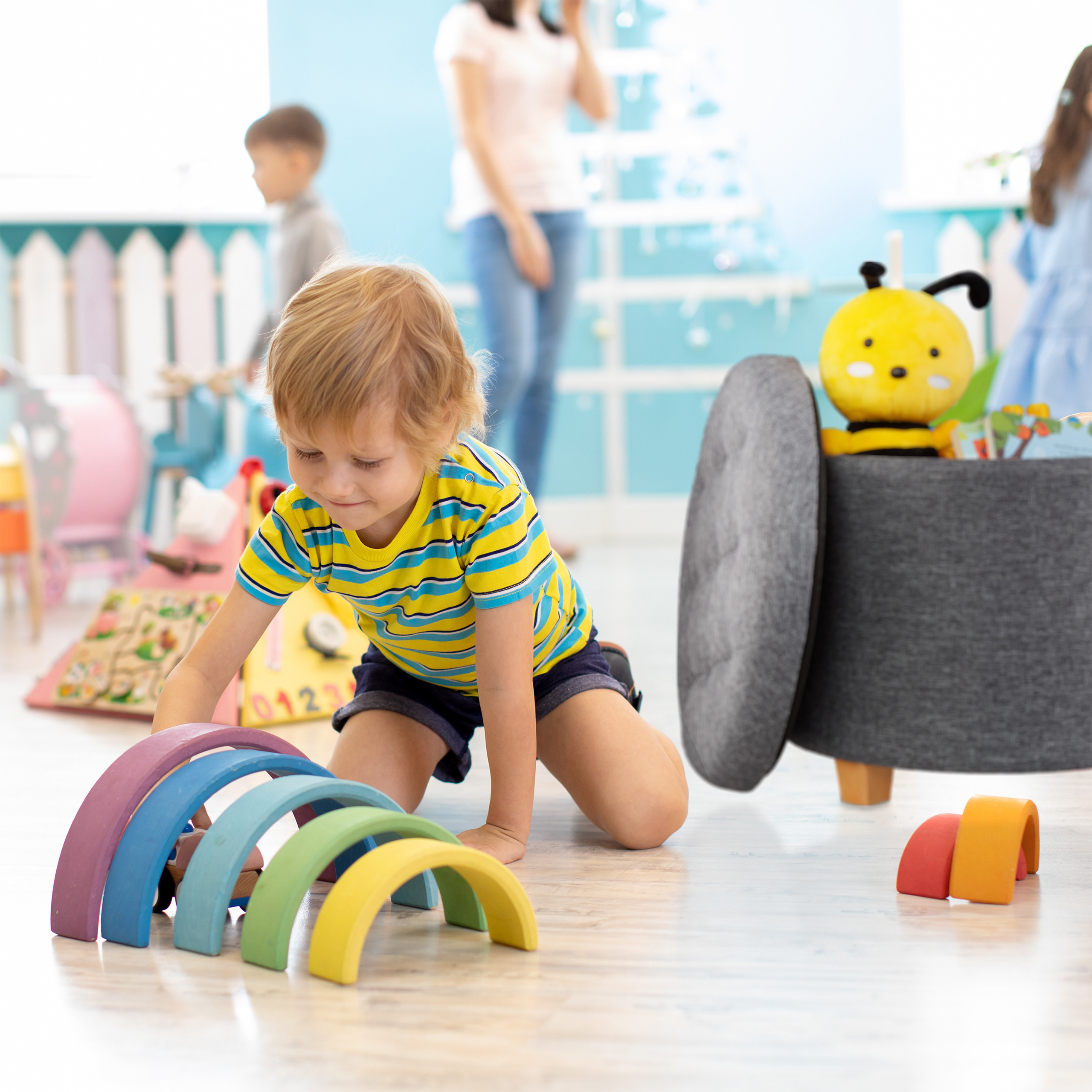 Kleinkind spielt auf dem Boden mit bunten Holzbögen, im Hintergrund sind andere Kinder und ein Spielzeugkorb.