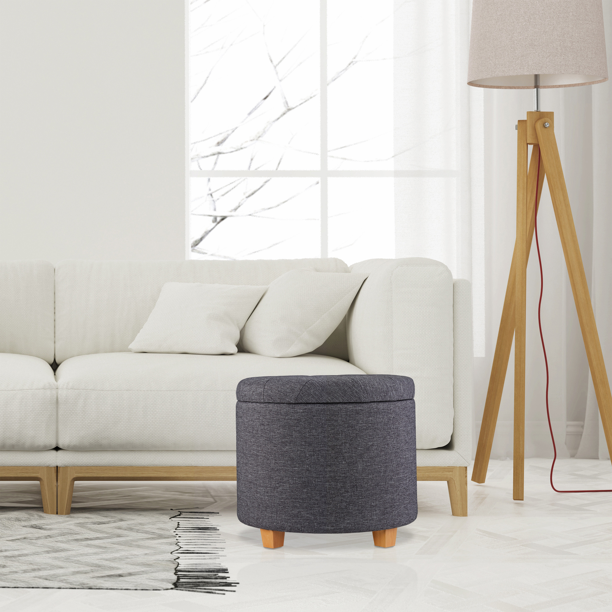 Ein grauer Stoffhocker mit Holzbeinen steht in einem hellen, modernen Wohnzimmer mit weißem Sofa und Stehlampe.