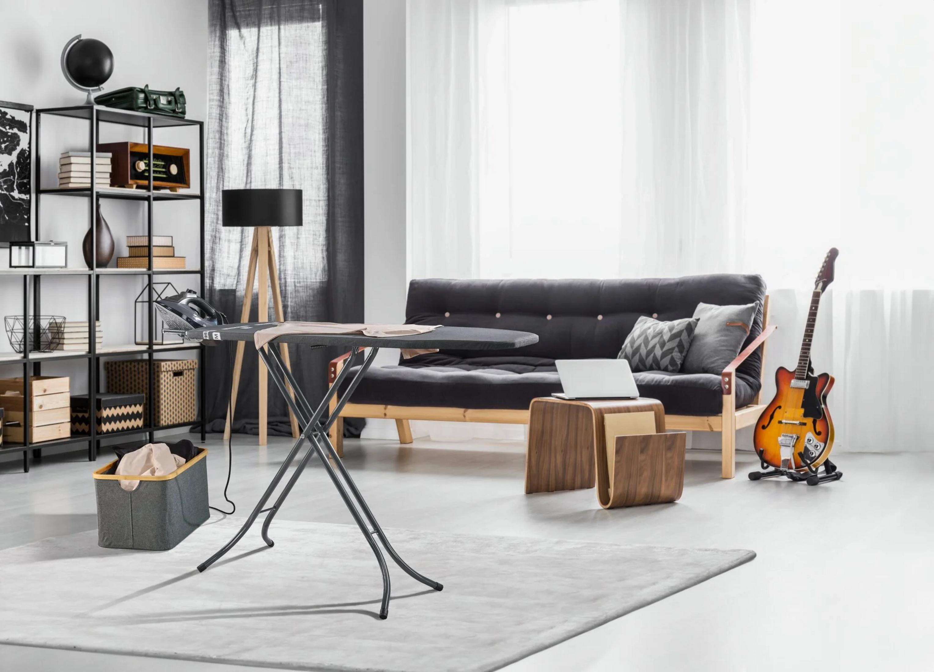 Wohnzimmer mit Bügelbrett im Vordergrund, schwarzem Sofa, Holzregal, Laptop und E-Gitarre.