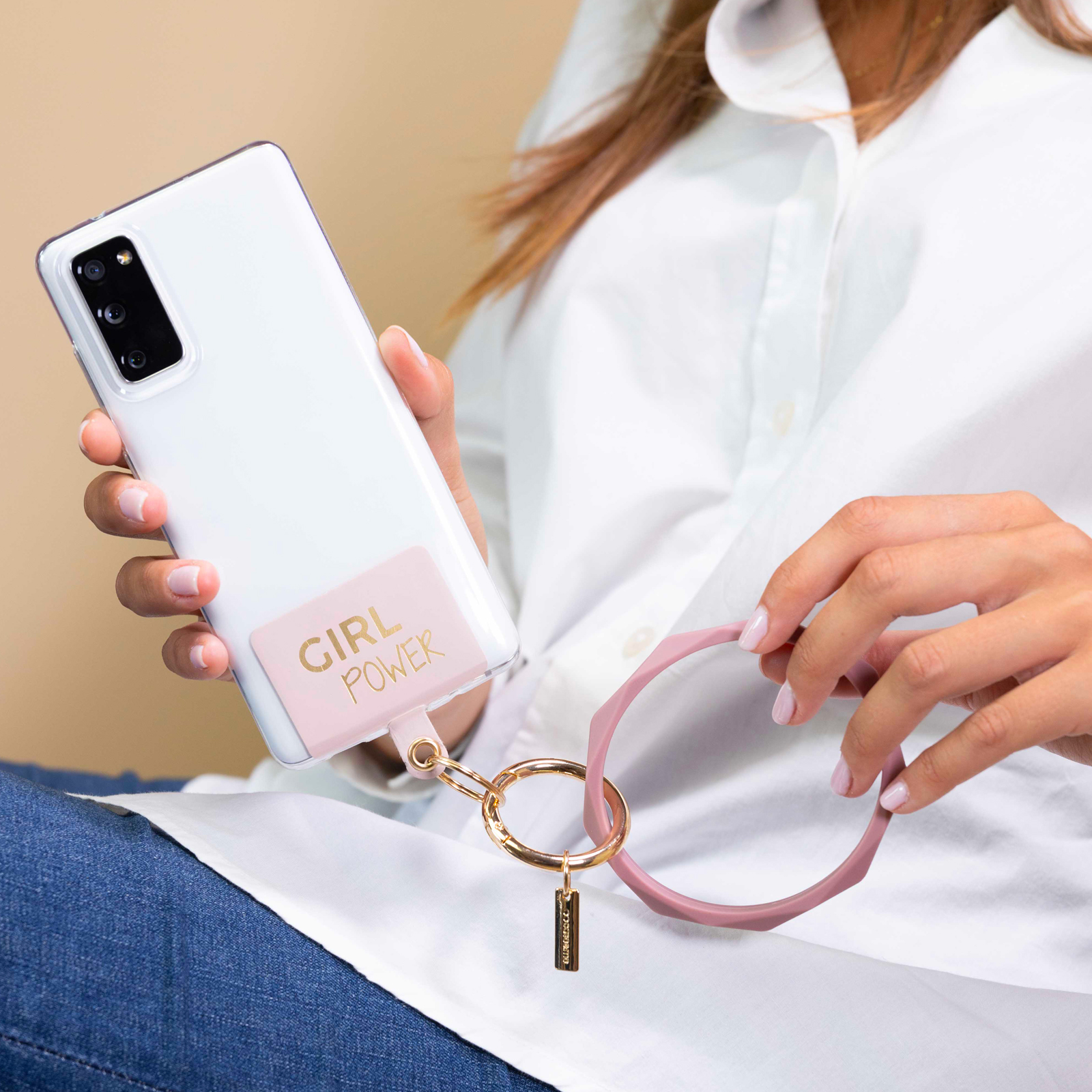 Una persona sostiene un smartphone blanco con correa rosa con texto 'GIRL POWER' y anillos dorados.