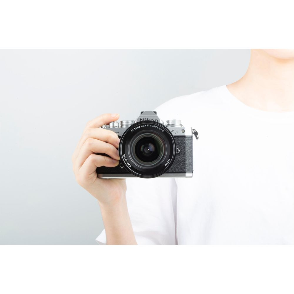 Persona con maglietta bianca tiene in mano una fotocamera mirrorless Nikon argento e nera con un obiettivo grande.