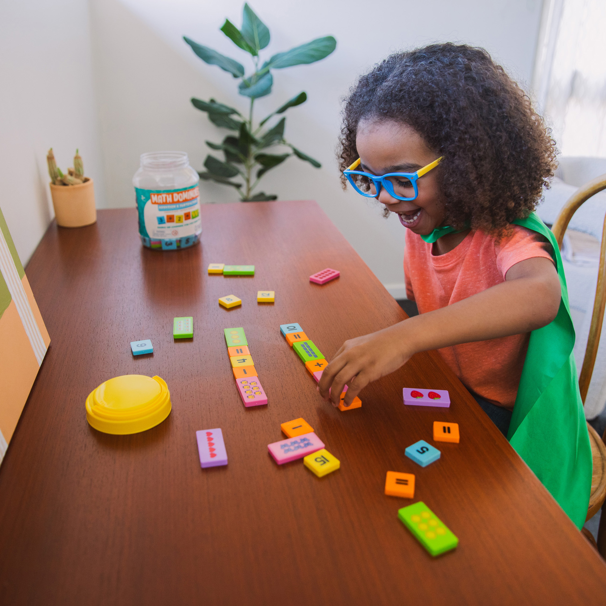 Ein Kind mit Brille und grünem Umhang spielt an einem Holztisch mit bunten Mathe-Domino-Steinen.