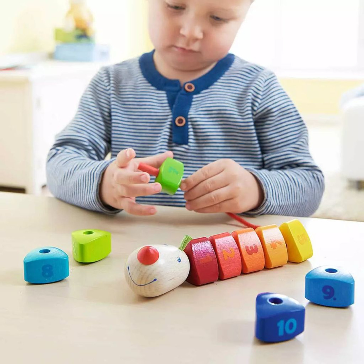 Kleinkind spielt am Tisch mit einem bunten hölzernen Raupen-Fädelspiel.