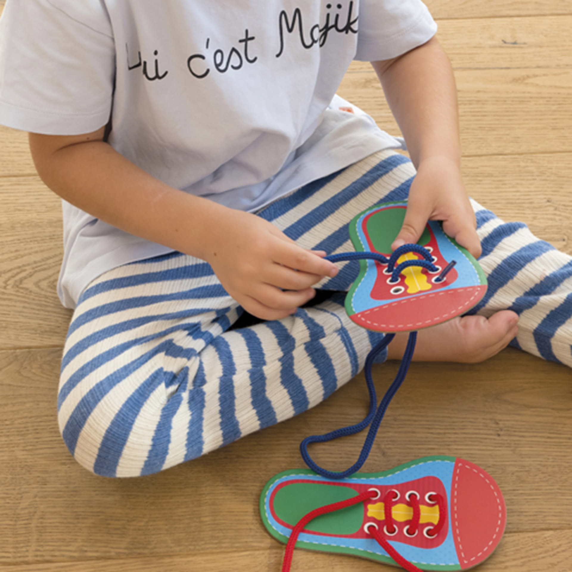 Un niño con pantalones de rayas se sienta en un suelo de madera, practicando lazada en un zapato de juguete.