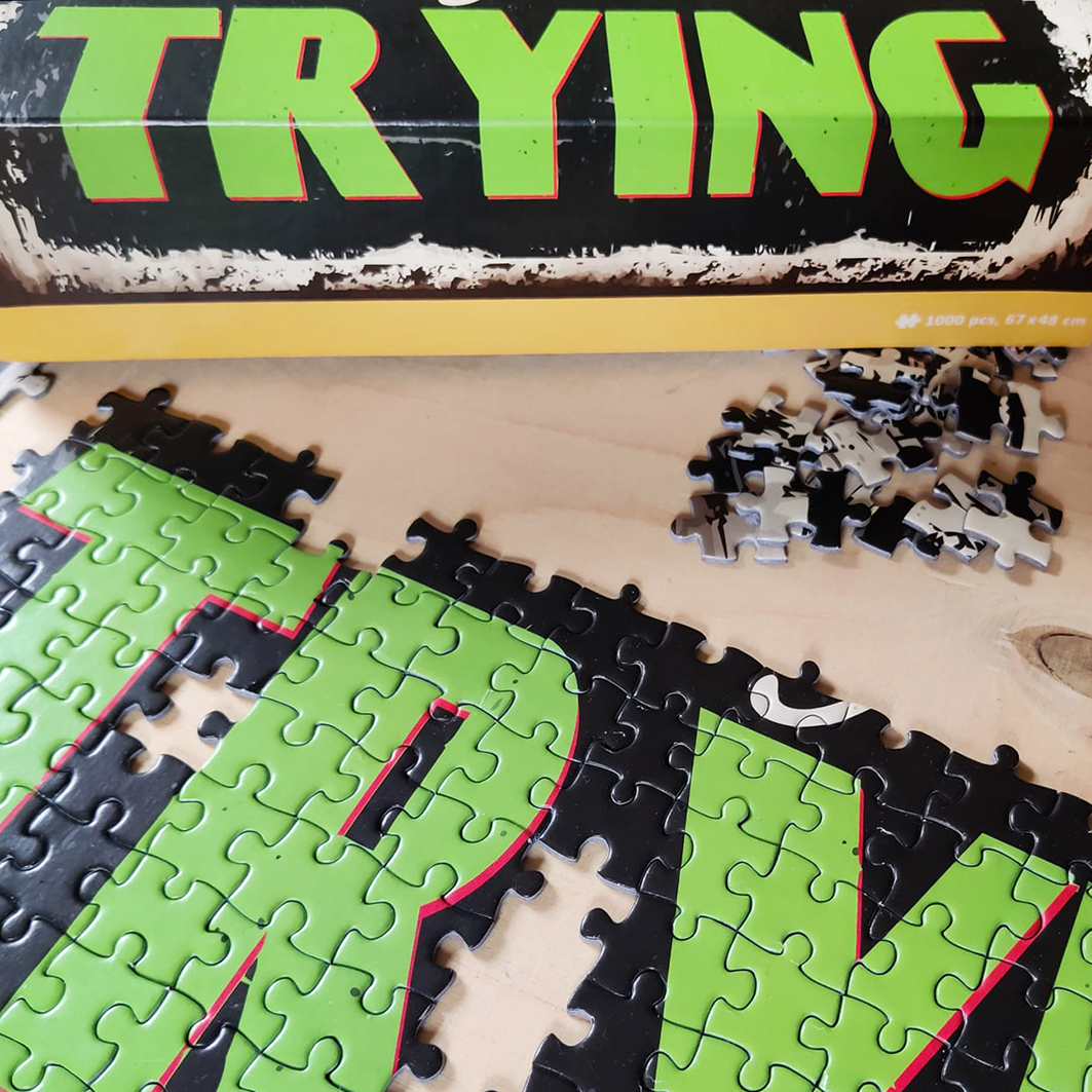 Ein unvollständiges Puzzle mit grünen und schwarzen Teilen auf einem Tisch, darunter der Text TRYING.