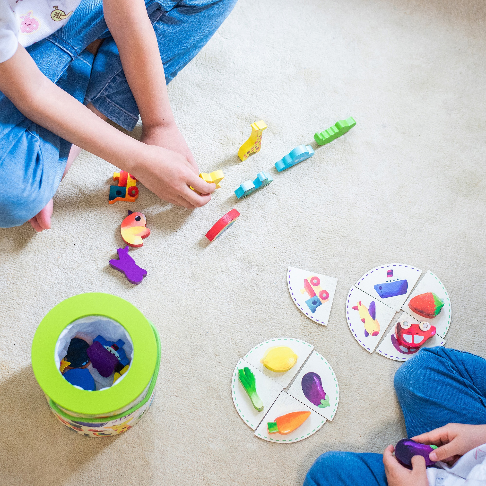 Zwei Kinder sitzen auf einem Teppich und spielen mit bunten Holzfiguren und runden Puzzleteilen.