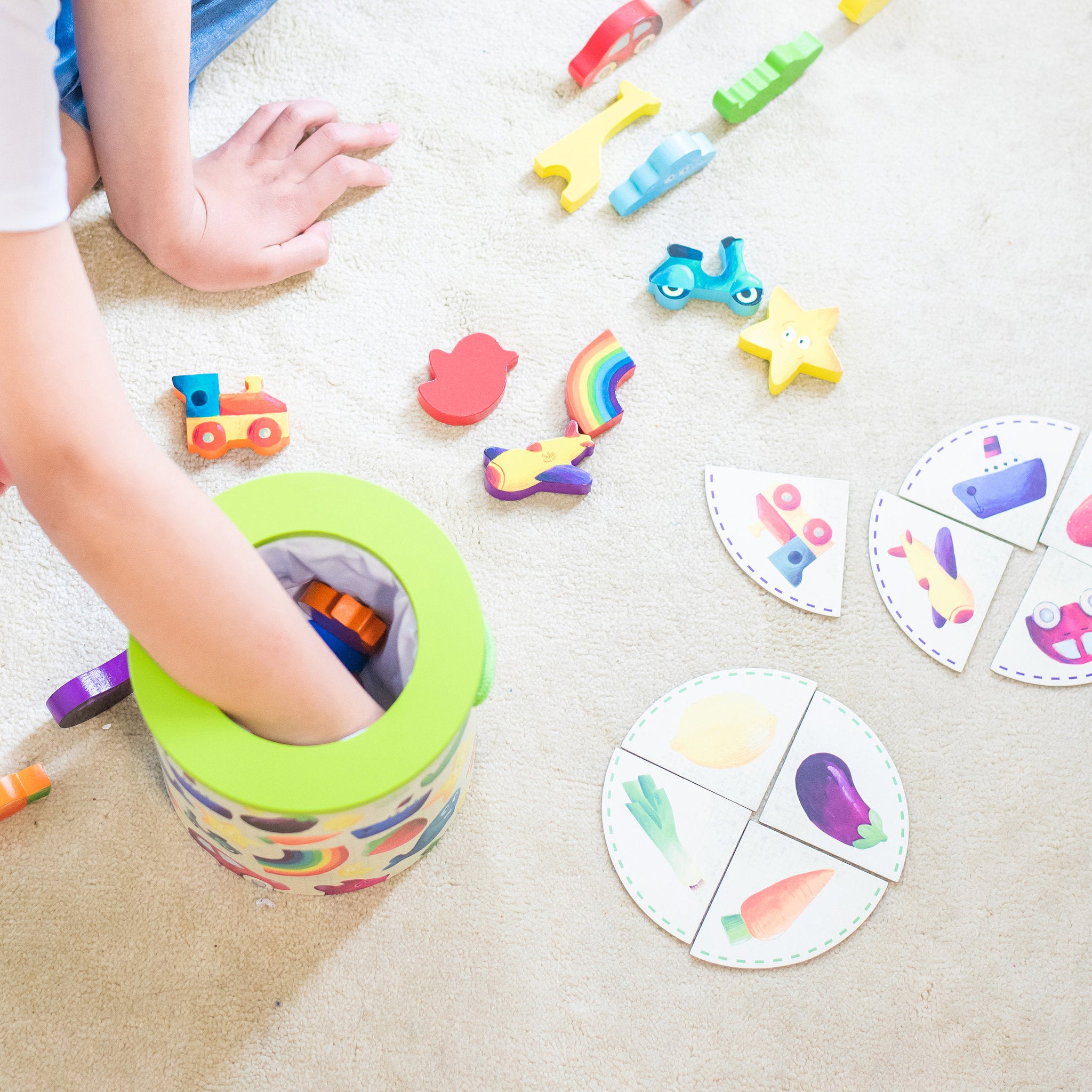 Ein Kind greift in eine bunte Spielzeugkiste, umgeben von Holzspielzeug und Puzzle-Teilen auf einem Teppich.