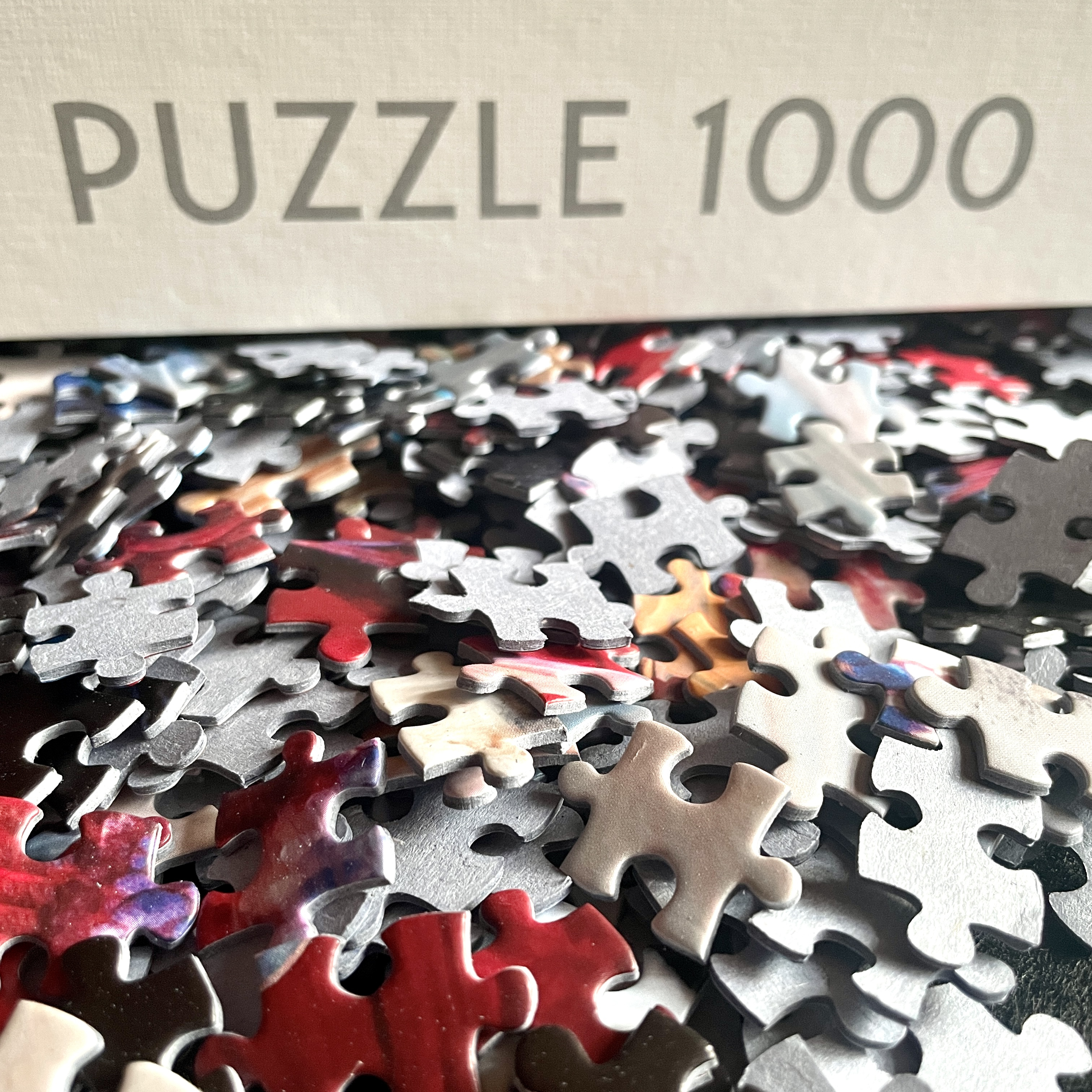 Un montón de piezas de rompecabezas coloridas frente a una caja con la inscripción 'PUZZLE 1000'.