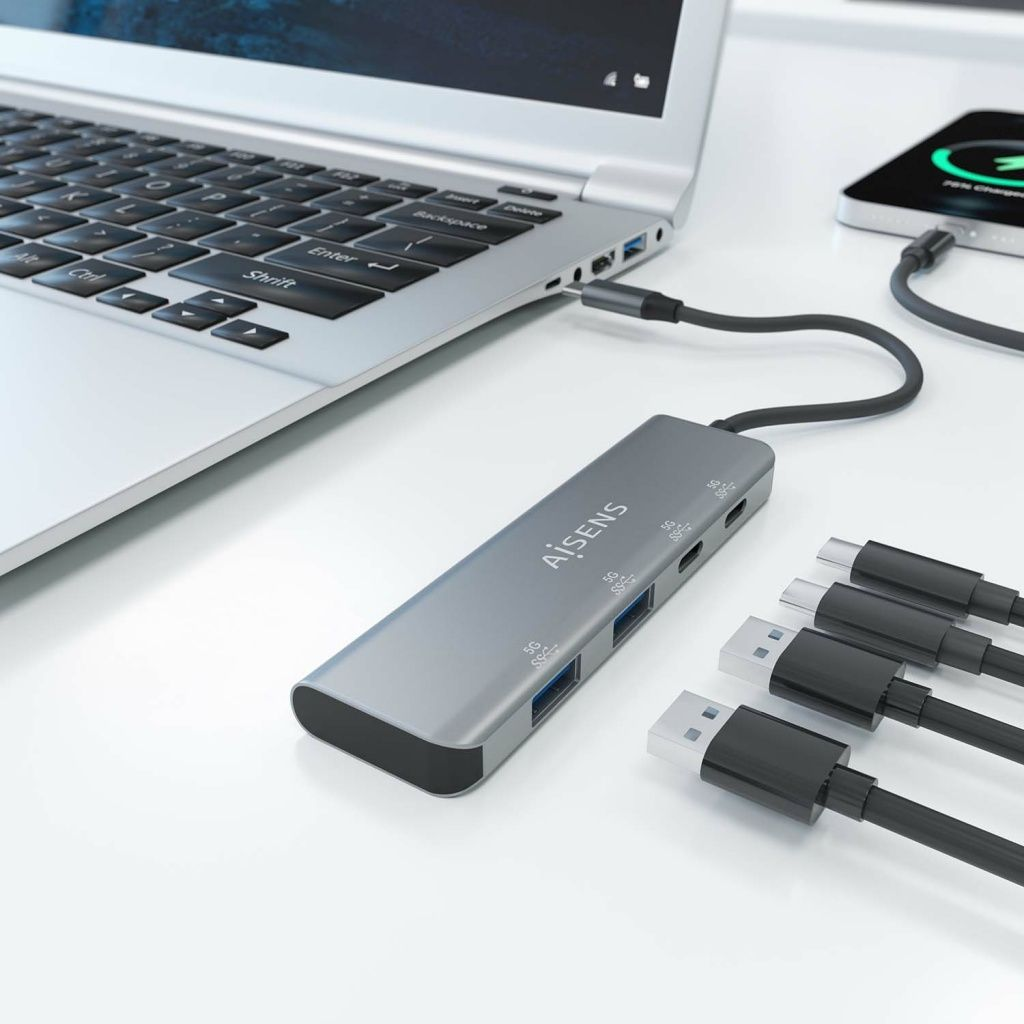 Hub USB-C gris con múltiples puertos conectado a una computadora portátil, con cables y un teléfono inteligente cerca.