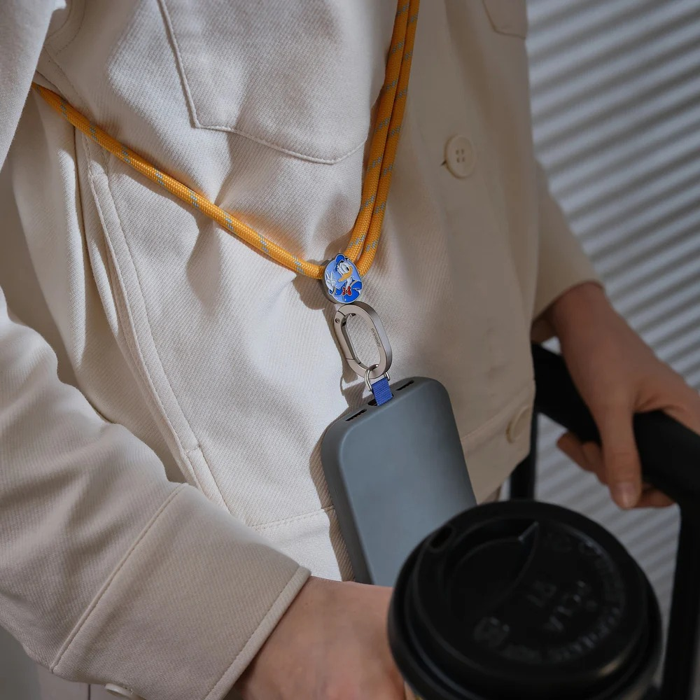 Osoba w białym stroju ma szary smartfon na żółtej smyczy i trzyma kubek kawy.