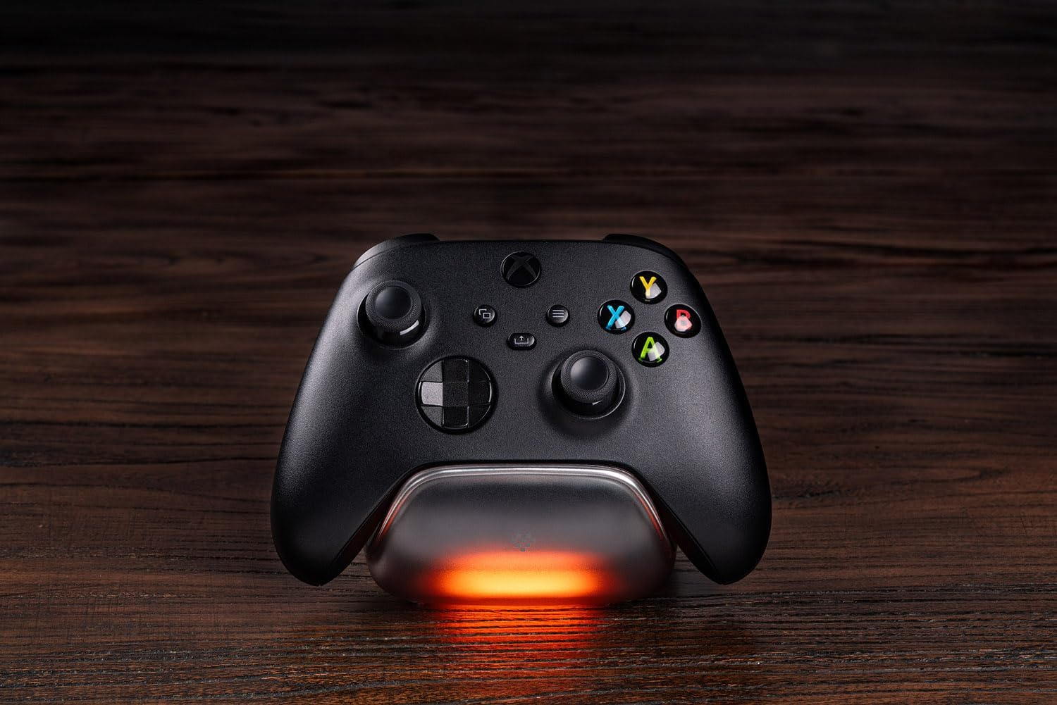 Czarny kontroler Xbox na świecącej pomarańczowej podstawce ładującej na ciemnym drewnie.