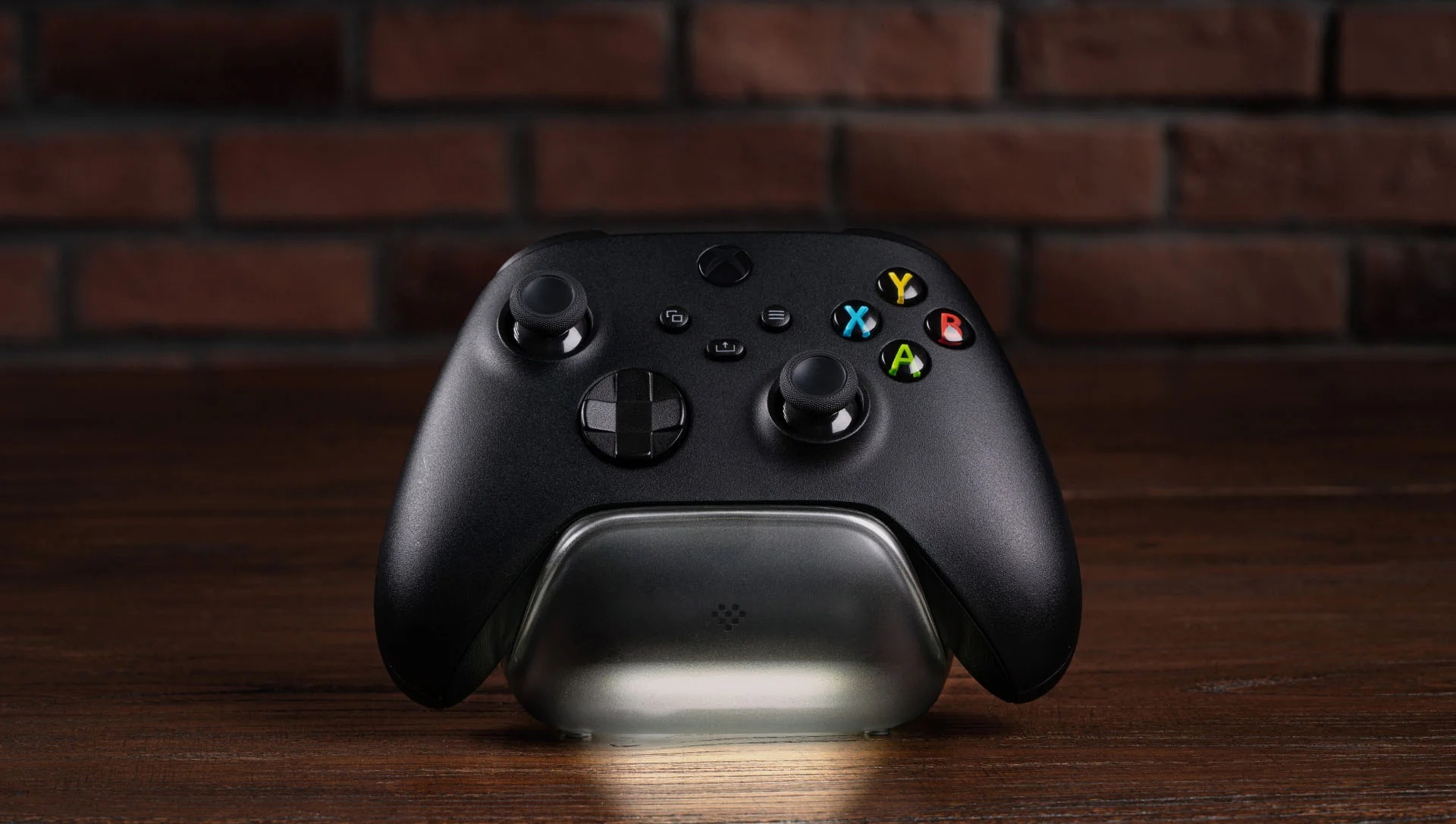 Czarny kontroler Xbox spoczywa na podświetlanej stacji ładującej przed ceglaną ścianą.