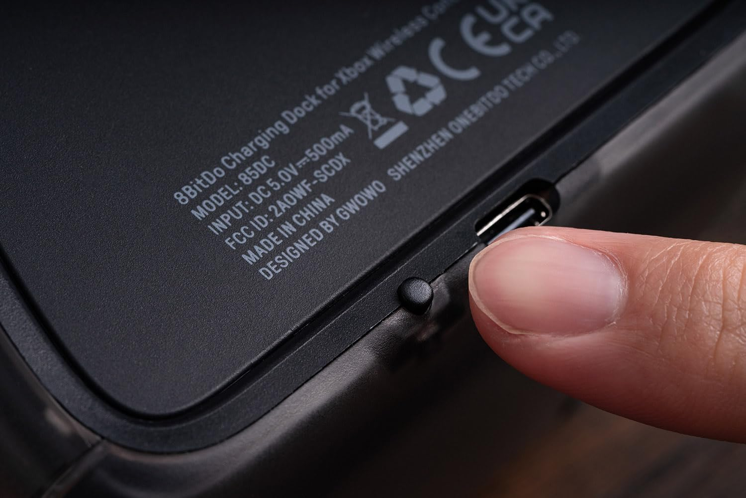 Palec dotyka małego przycisku obok portu USB-C na czarnym urządzeniu z białym nadrukiem.