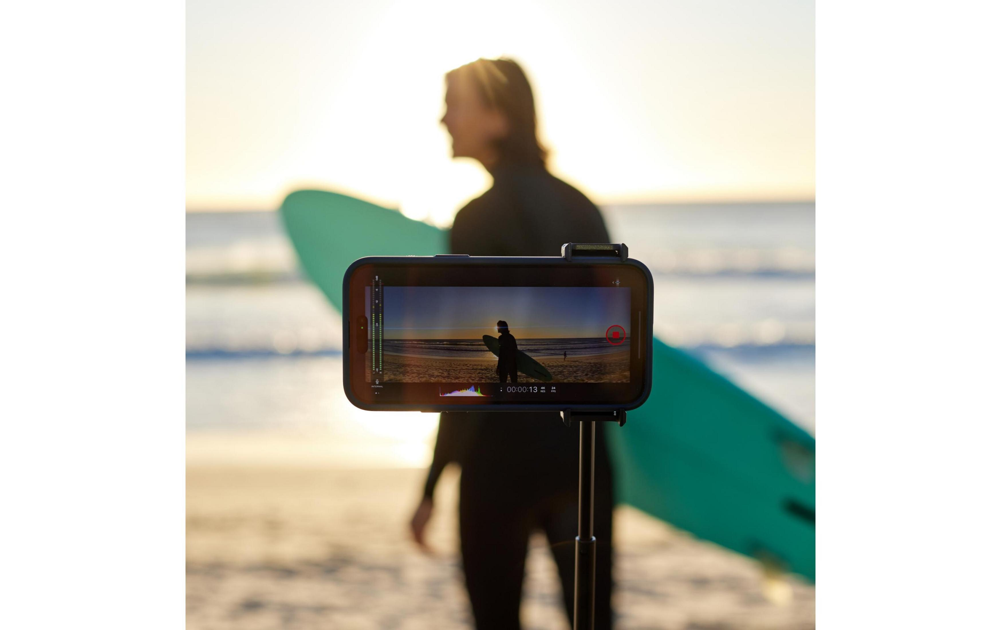 Smartfon na statywie nagrywa surfer trzymającego turkusową deskę na plaży o zachodzie słońca.