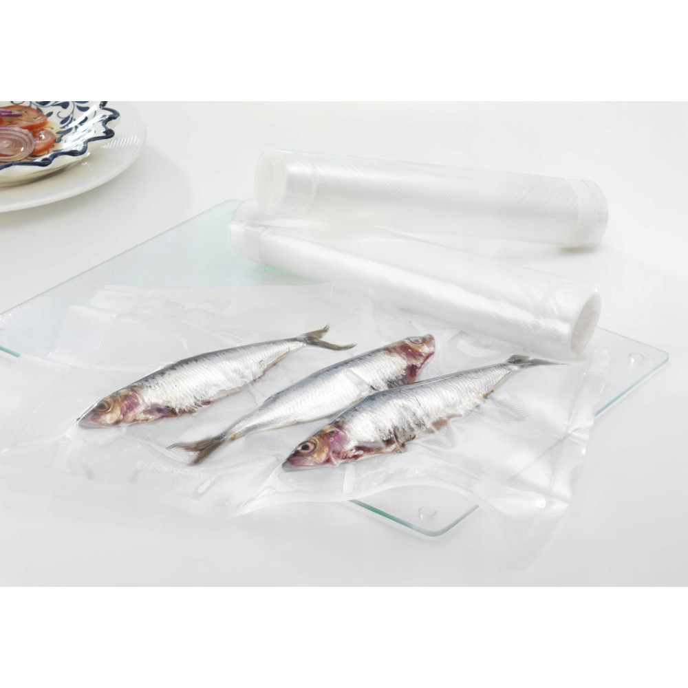 Tres sardinas frescas en bolsa de vacío sobre tabla de vidrio, con rollos de film.