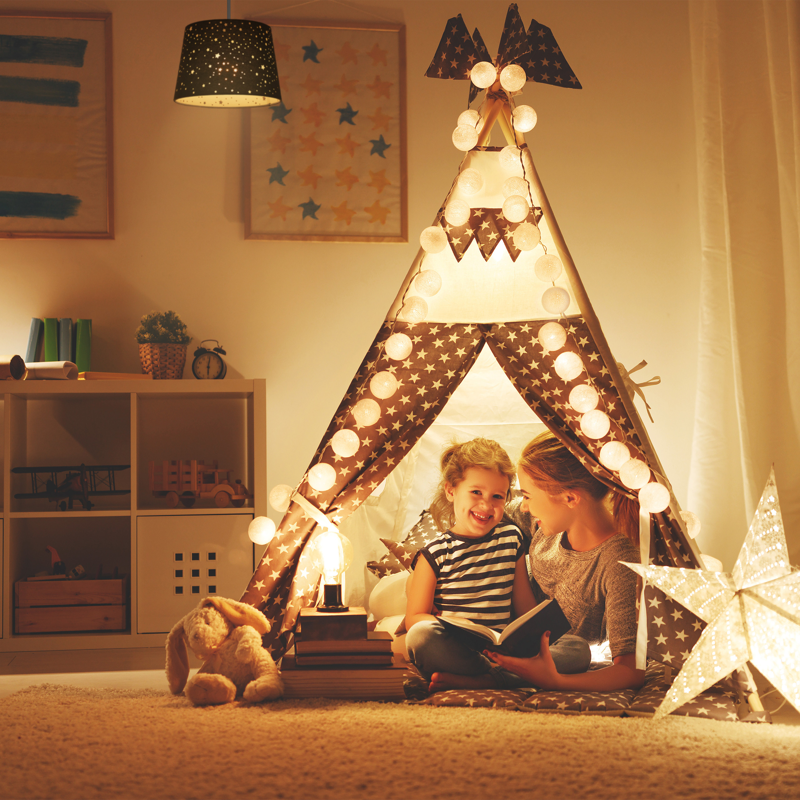 Mutter und Tochter lesen zusammen ein Buch in einem beleuchteten Tipi-Zelt im Kinderzimmer.