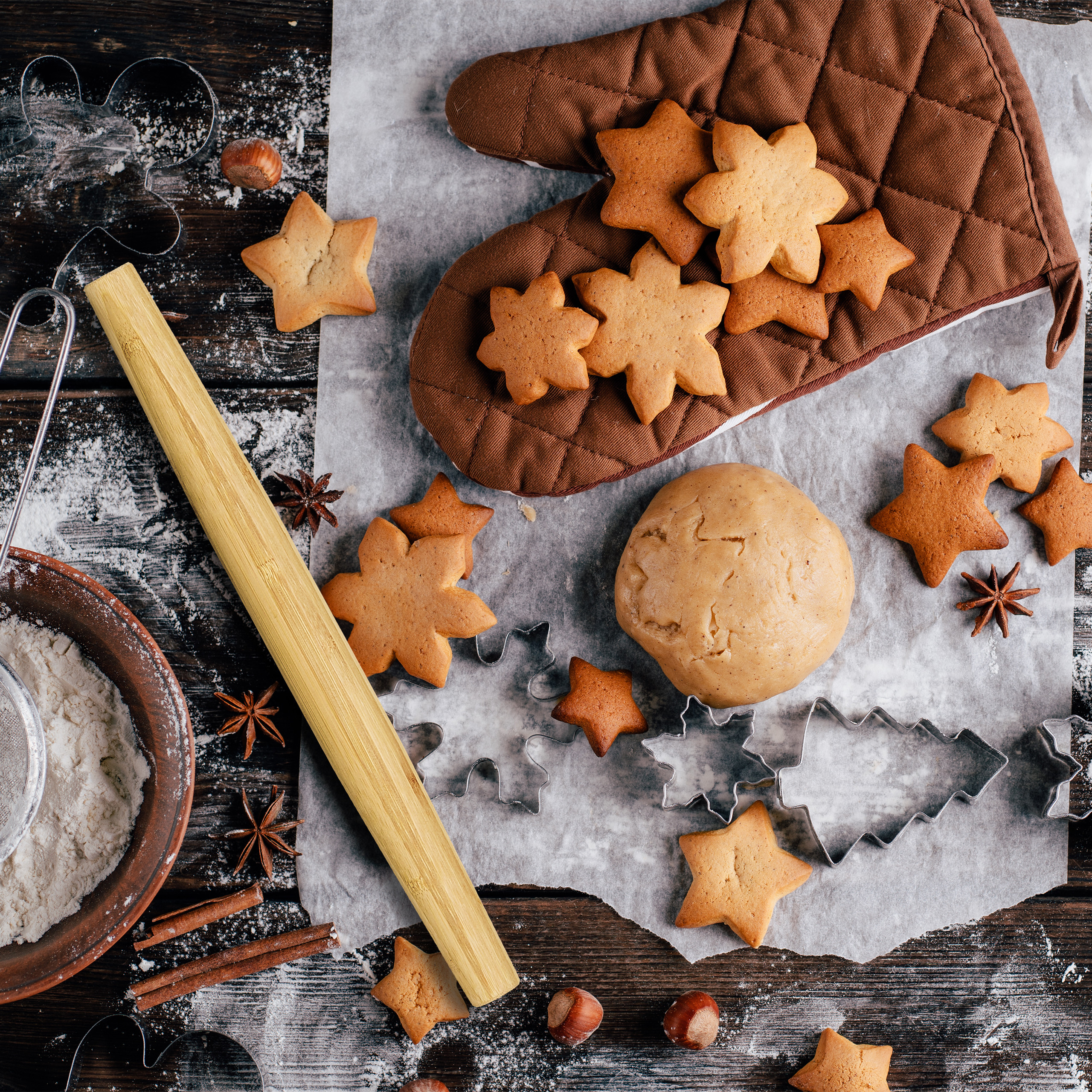 Backszene mit Sternplätzchen, Teigkugel, Nudelholz, Ofenhandschuh, Mehl und Sternanis auf dunklem Holztisch.