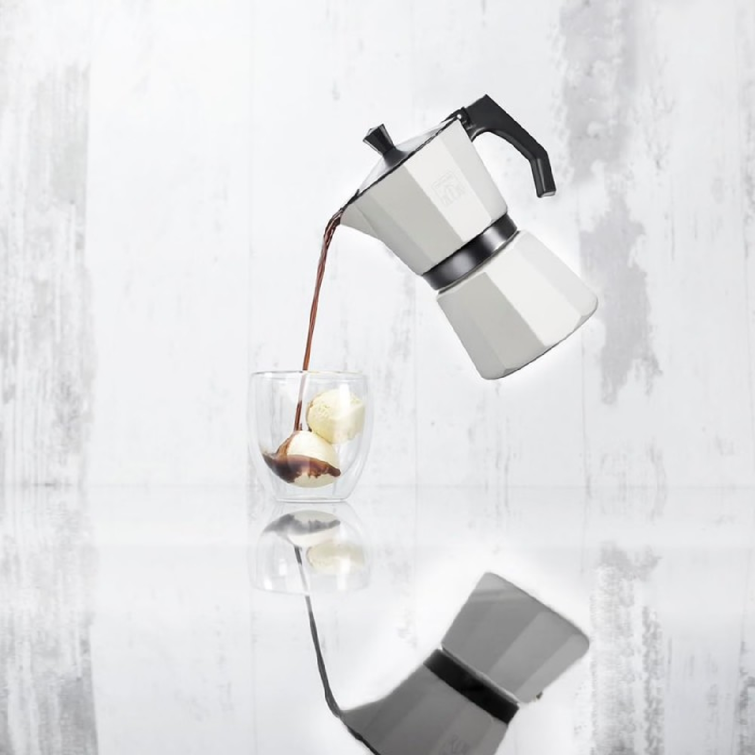 Una moka bianca versa caffè in un bicchiere con palline di gelato su sfondo chiaro.