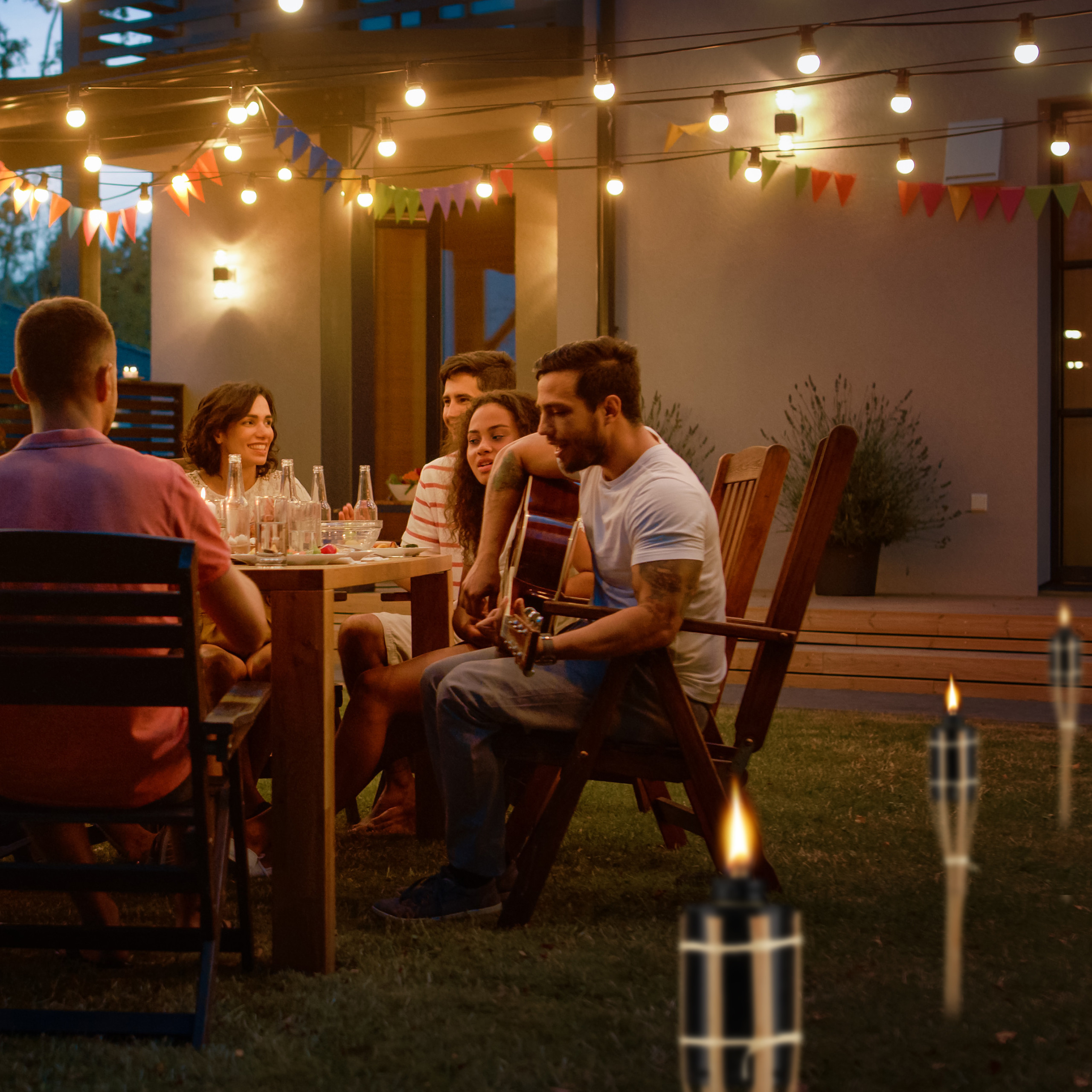 Eine Gruppe junger Freunde sitzt abends im Garten an einem Holztisch, beleuchtet von Lichterketten und Fackeln.