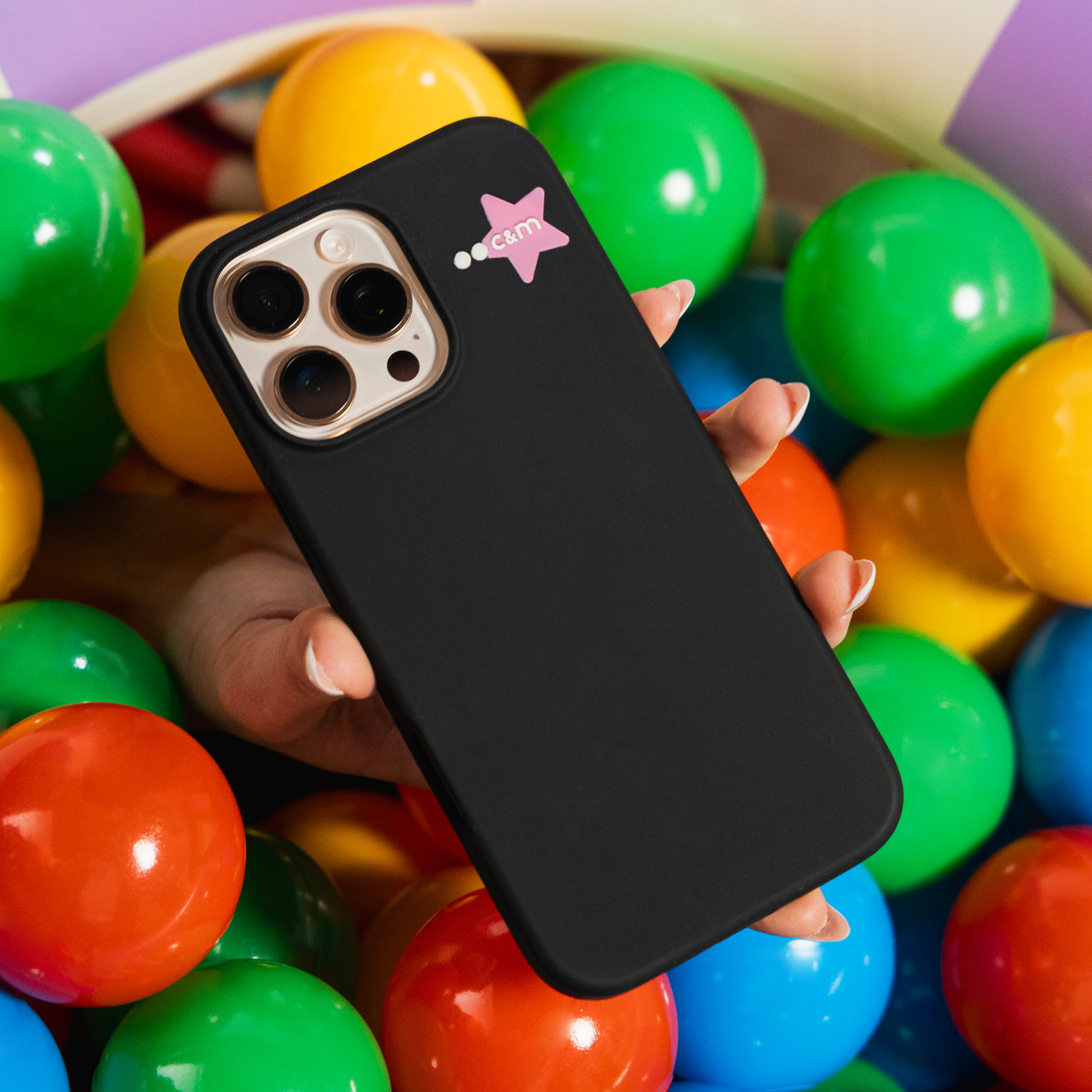 Una mano sosteniendo una funda de teléfono negra con una pegatina de estrella rosa, rodeada de pelotas de plástico de colores.