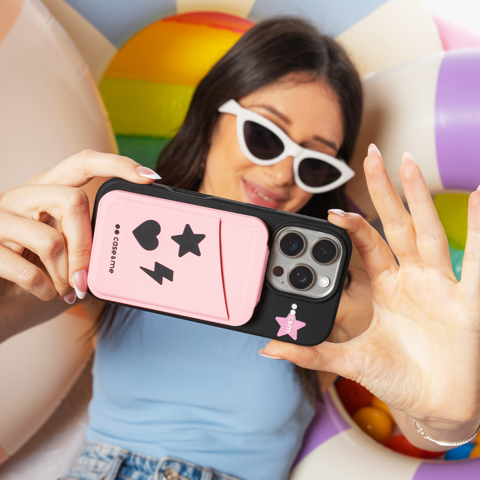 Una mujer con gafas de sol blancas se hace un selfie con una funda rosa y negra ante aros de colores.