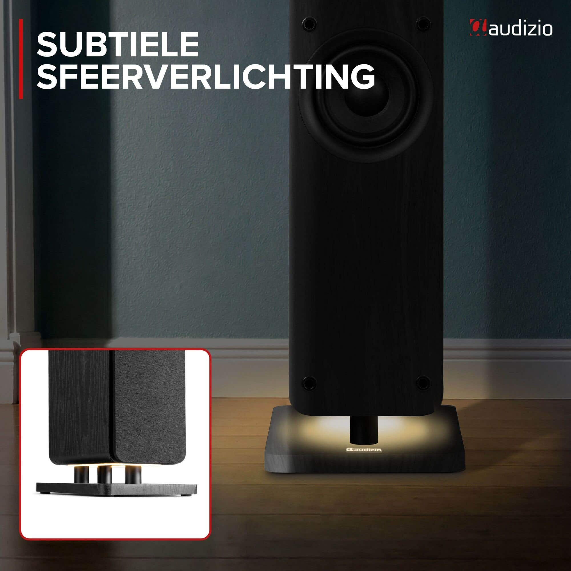 Un altavoz de torre negro sobre suelo de madera con suave luz base, con un primer plano de sus patas.