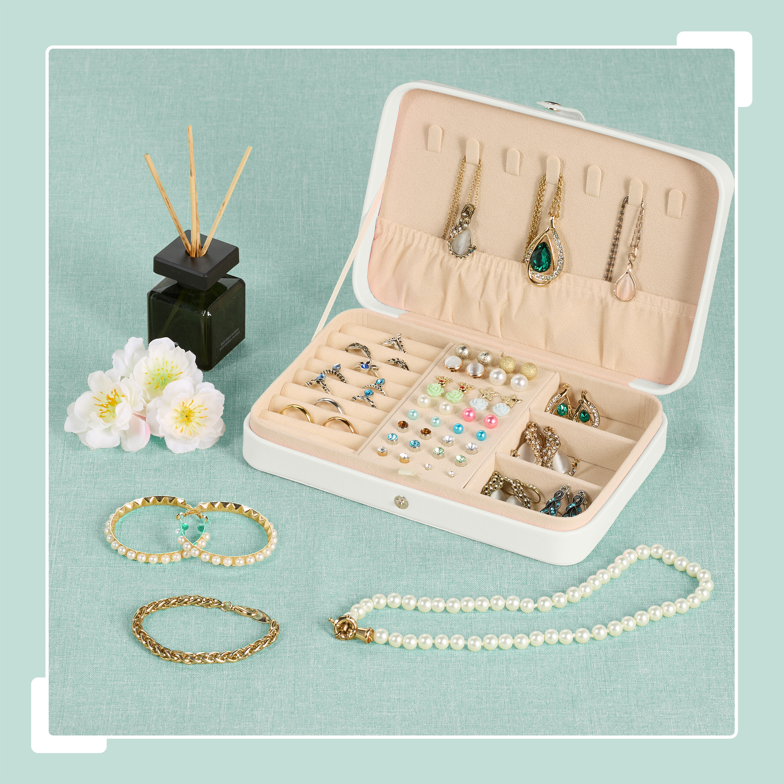 Eine weiße Schmuckschatulle gefüllt mit Ringen, Ohrringen und Ketten, umgeben von Armbändern, Blumen und einem Duftflakon.