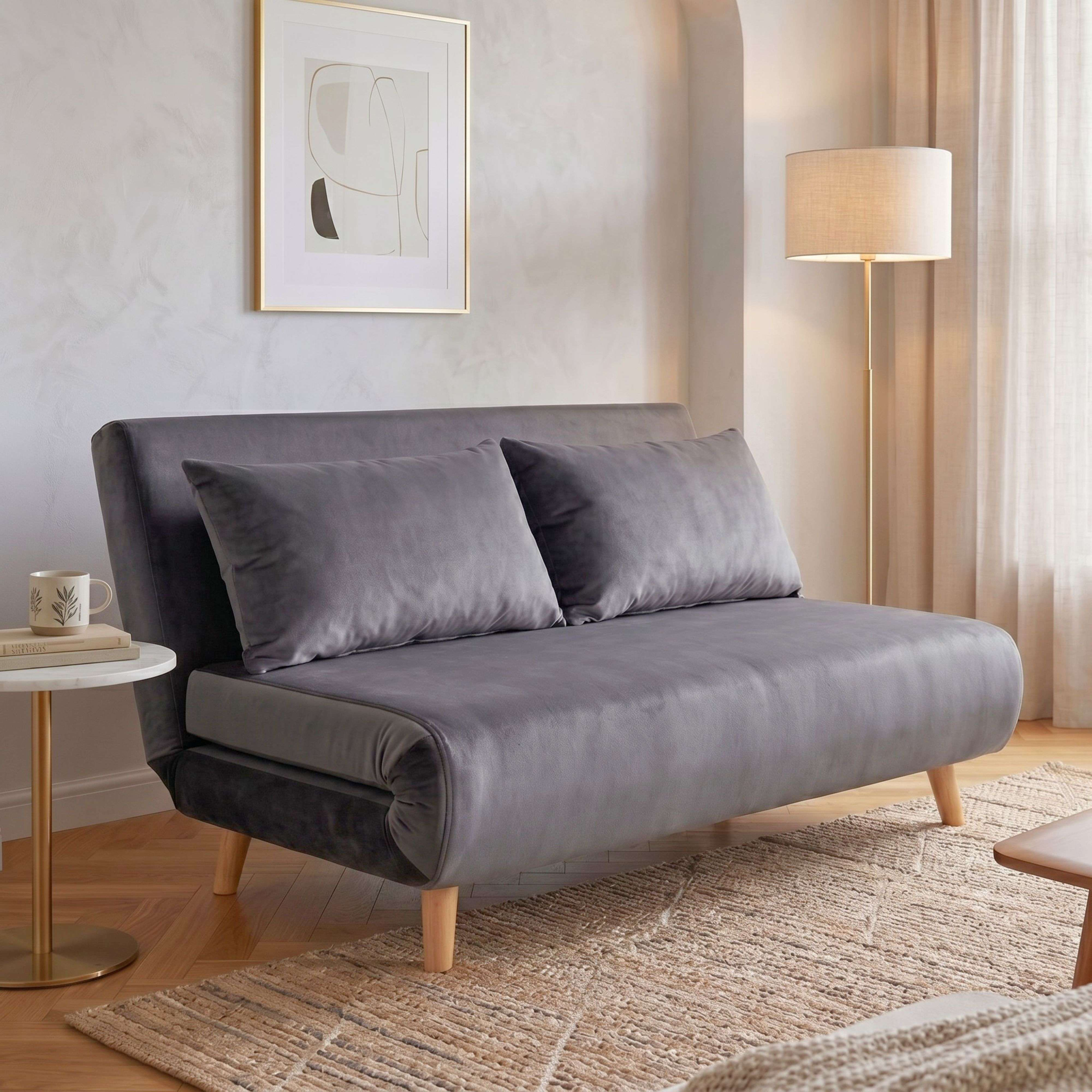 Un sofá cama gris con cojines en una sala luminosa junto a una lámpara de pie y una mesa auxiliar redonda.