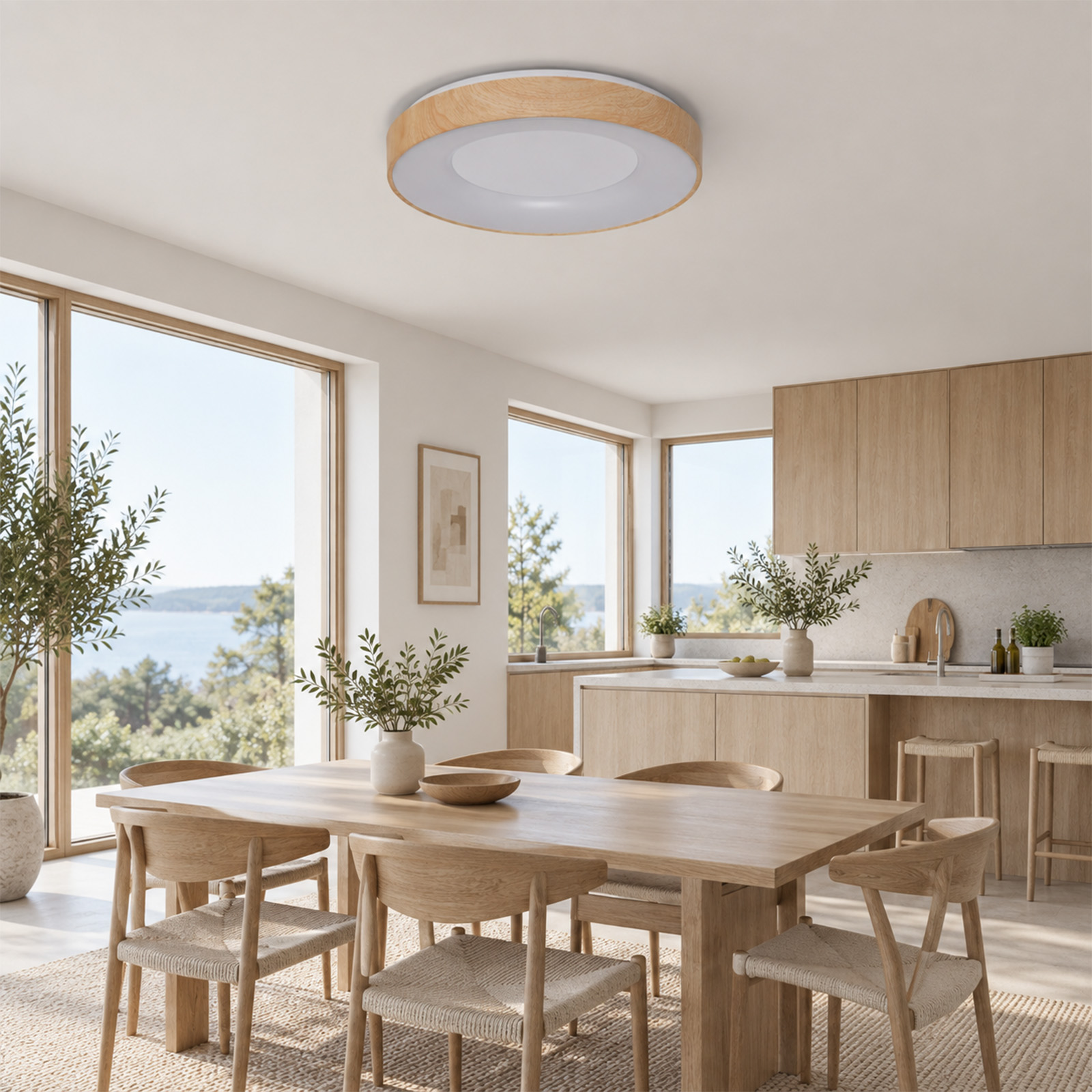 Área de comedor y cocina luminosa con muebles de madera clara, grandes ventanas con vista al agua y lámpara de techo.