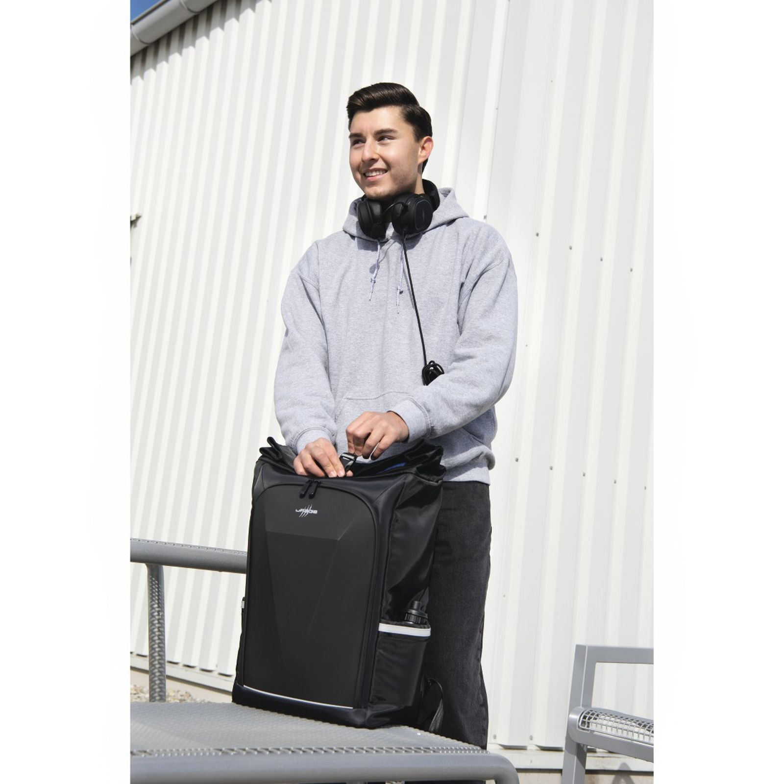Ein junger Mann mit grauen Hoodie steht draußen vor einer weißen Wand und öffnet einen schwarzen Rucksack auf einem Tisch.