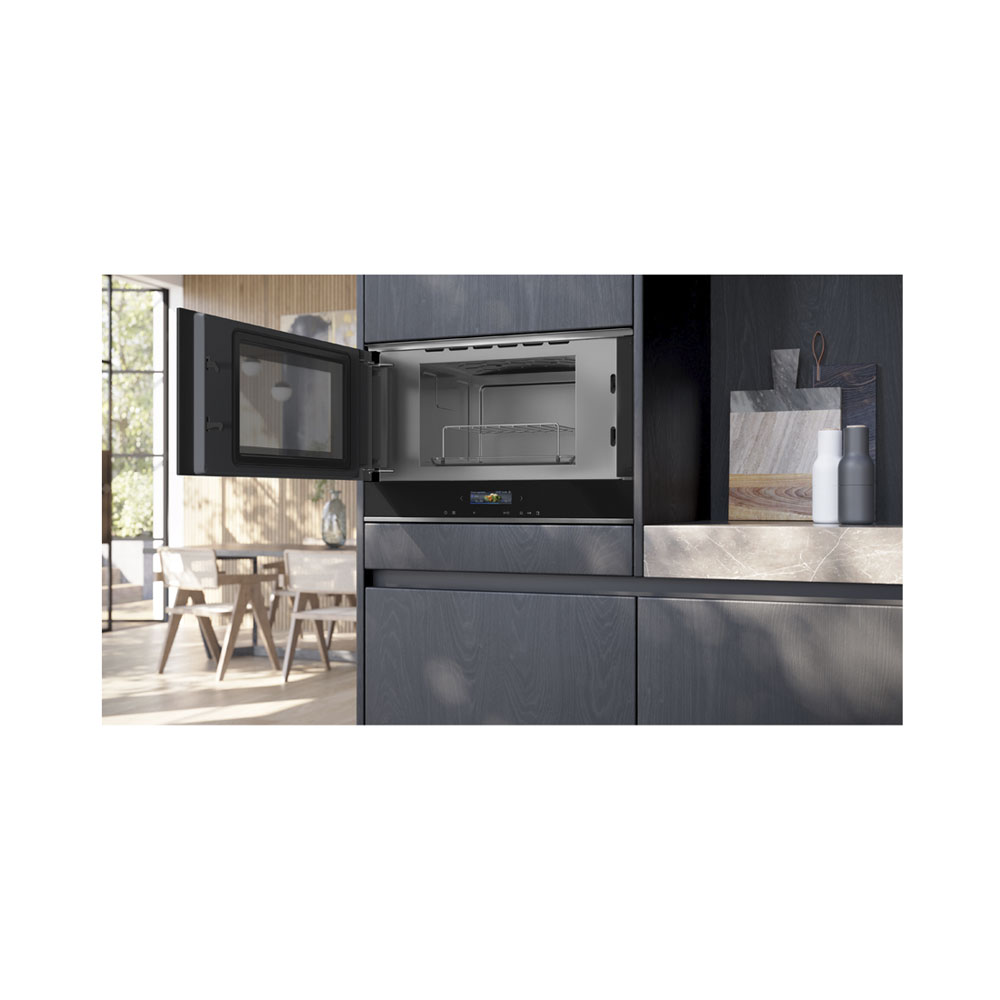 Un moderno forno incassato nero con la porta aperta in una cucina con mobili in legno e zona pranzo sullo sfondo.