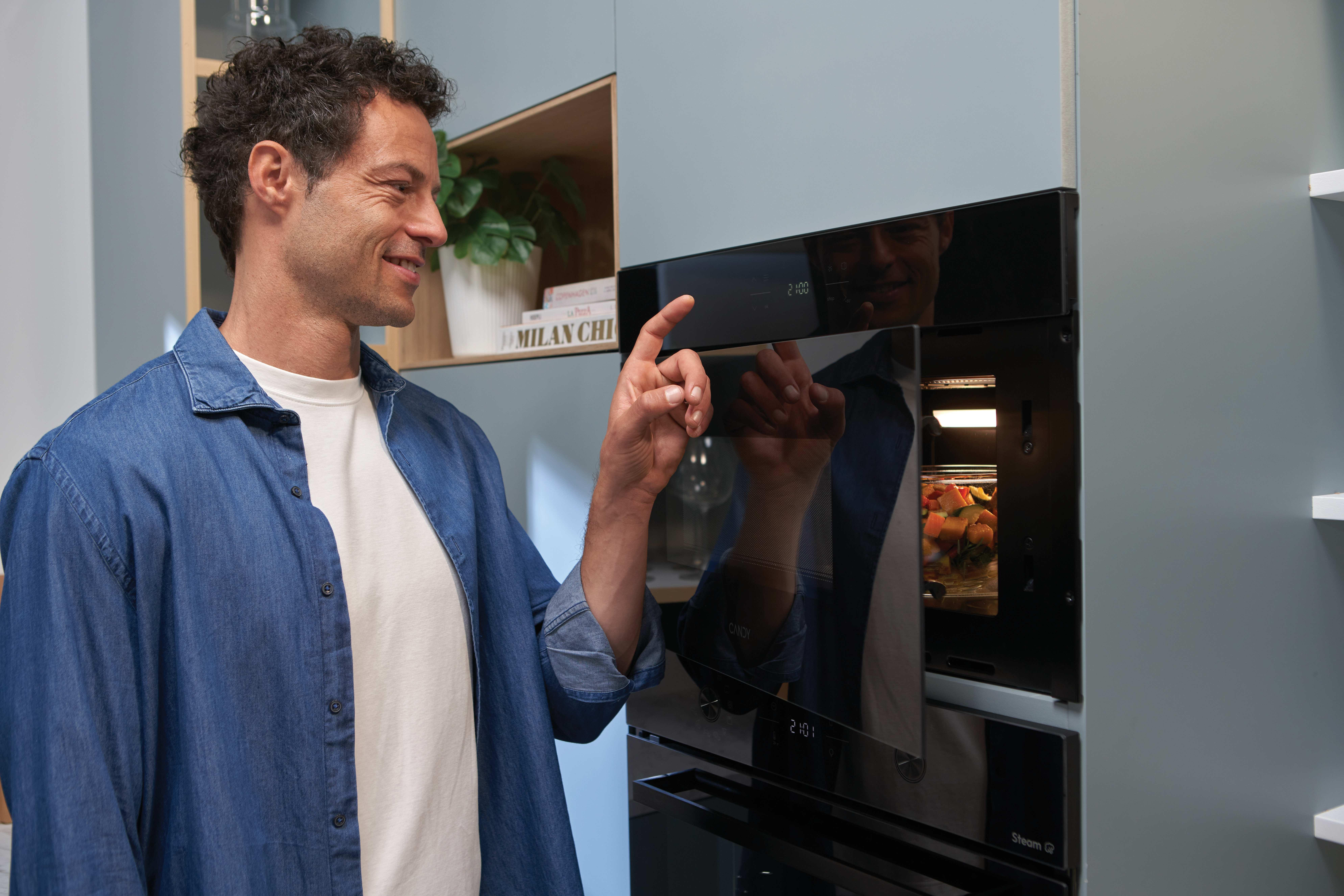 Un uomo sorridente con camicia di jeans tocca il pannello di controllo di un forno da incasso in cucina.