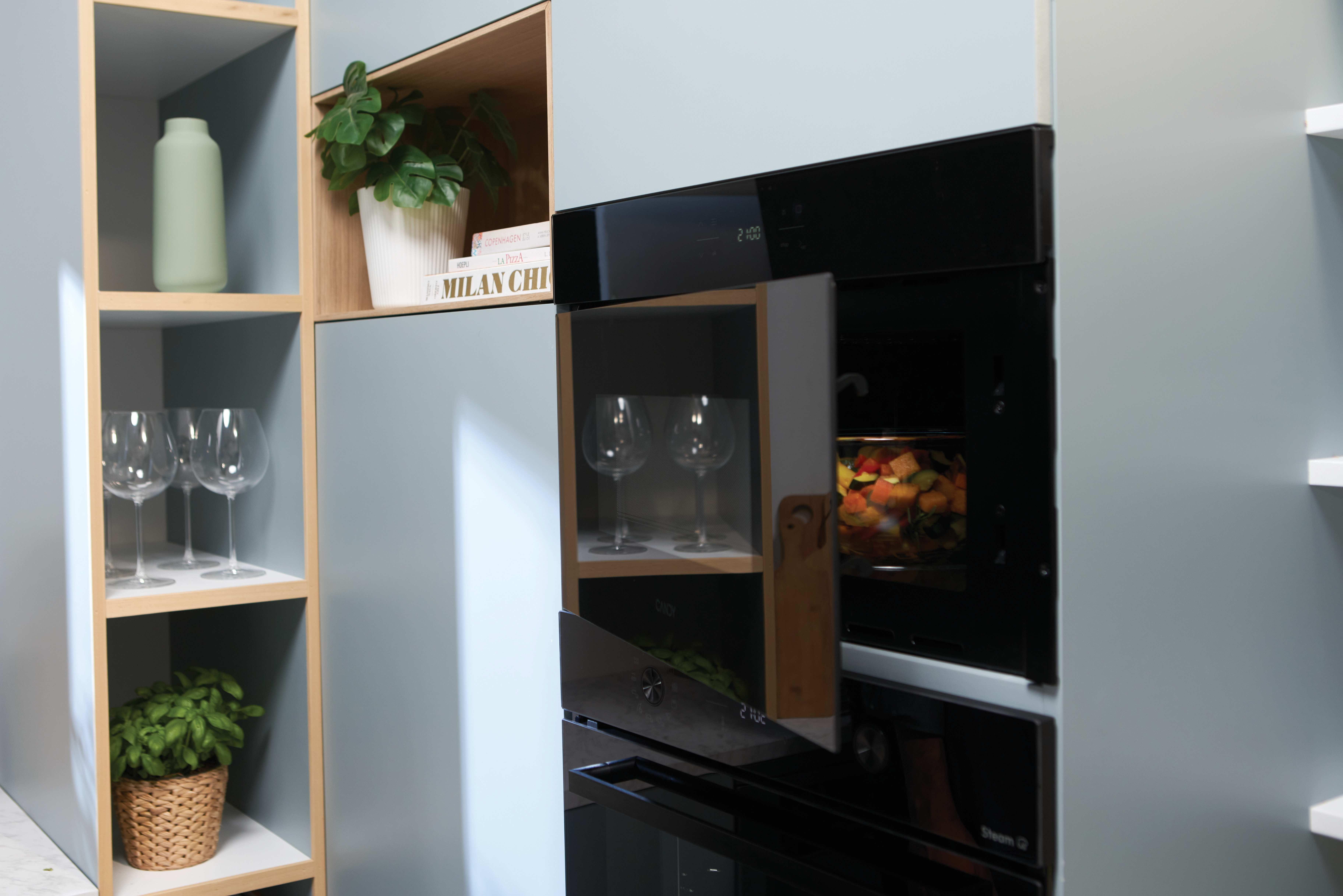 Una cucina moderna con forno incassato con verdure, ripiani con bicchieri e piante.