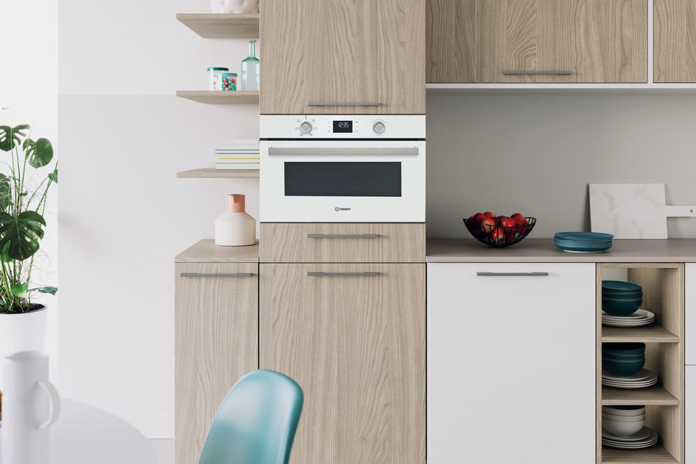 Una cucina moderna ha mobili in legno chiaro, un forno bianco da incasso, mensole con decorazioni e mele rosse.