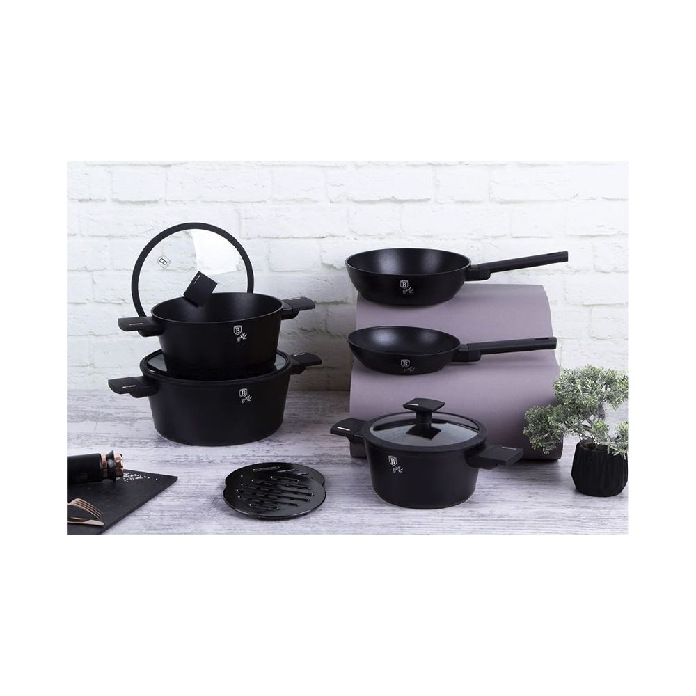Un ensemble de casseroles et poêles noires avec couvercles en verre sur une table devant un mur de briques blanches.