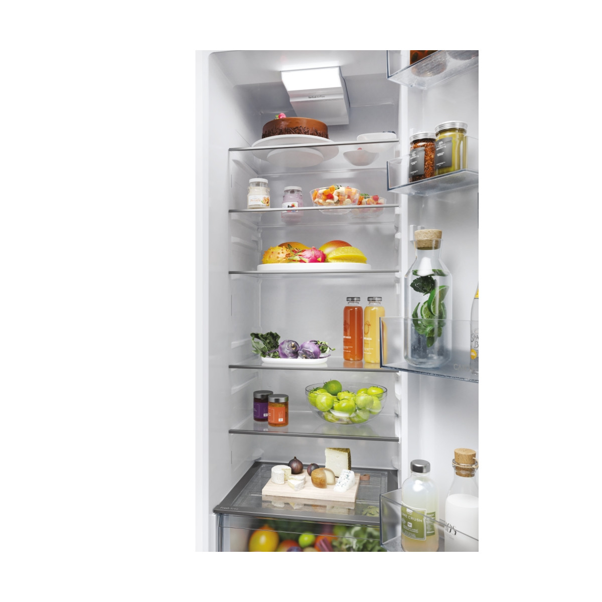 Un refrigerador blanco con estantes iluminados llenos de frascos, fruta fresca, verduras, bebidas y una tabla de quesos.